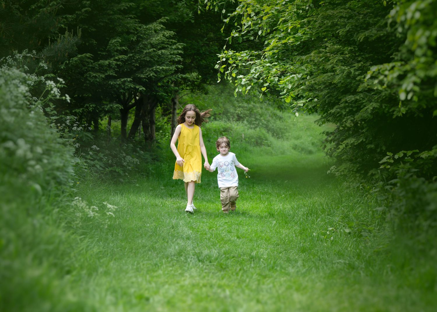 Mother and daughter holding hands while walking through a vibrant green path surrounded by tall trees and grass, both dressed in pastel colo