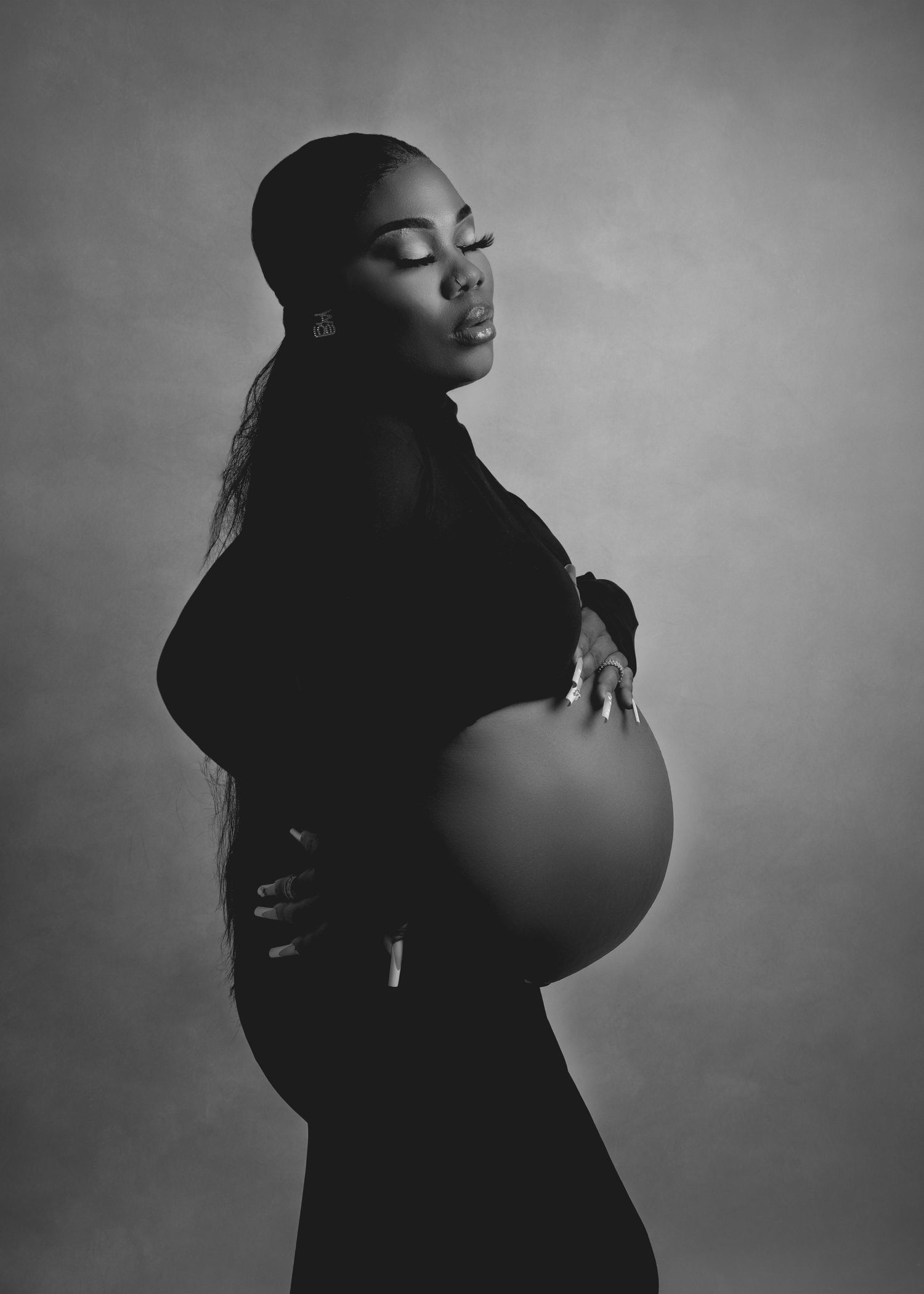 Black and white profile shot of a pregnant woman in a dark outfit, cradling her belly with a soft, thoughtful expression.