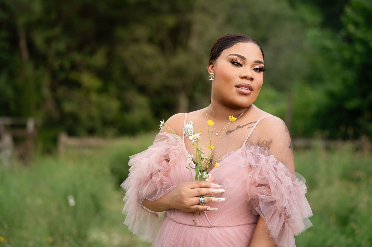 Woman standing outdoors in a lush green setting, wearing a pink sheer dress with floral details and holding a single flower, looking confide