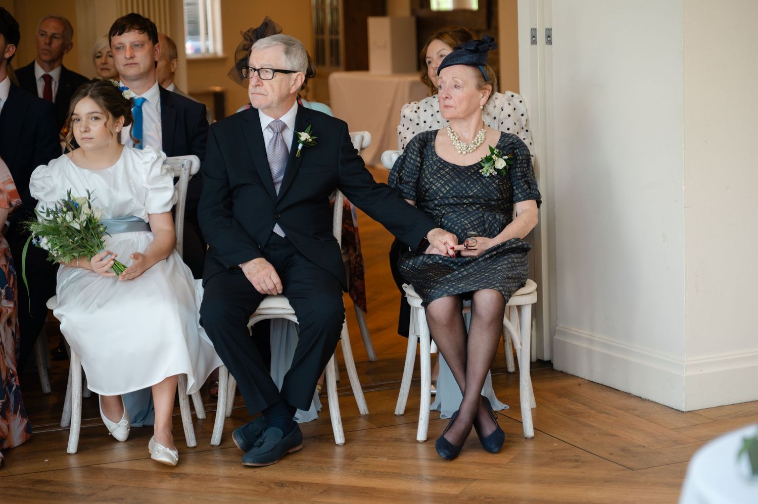 Guests seated and dressed formally, watching the wedding ceremony indoors with natural light coming through windows.