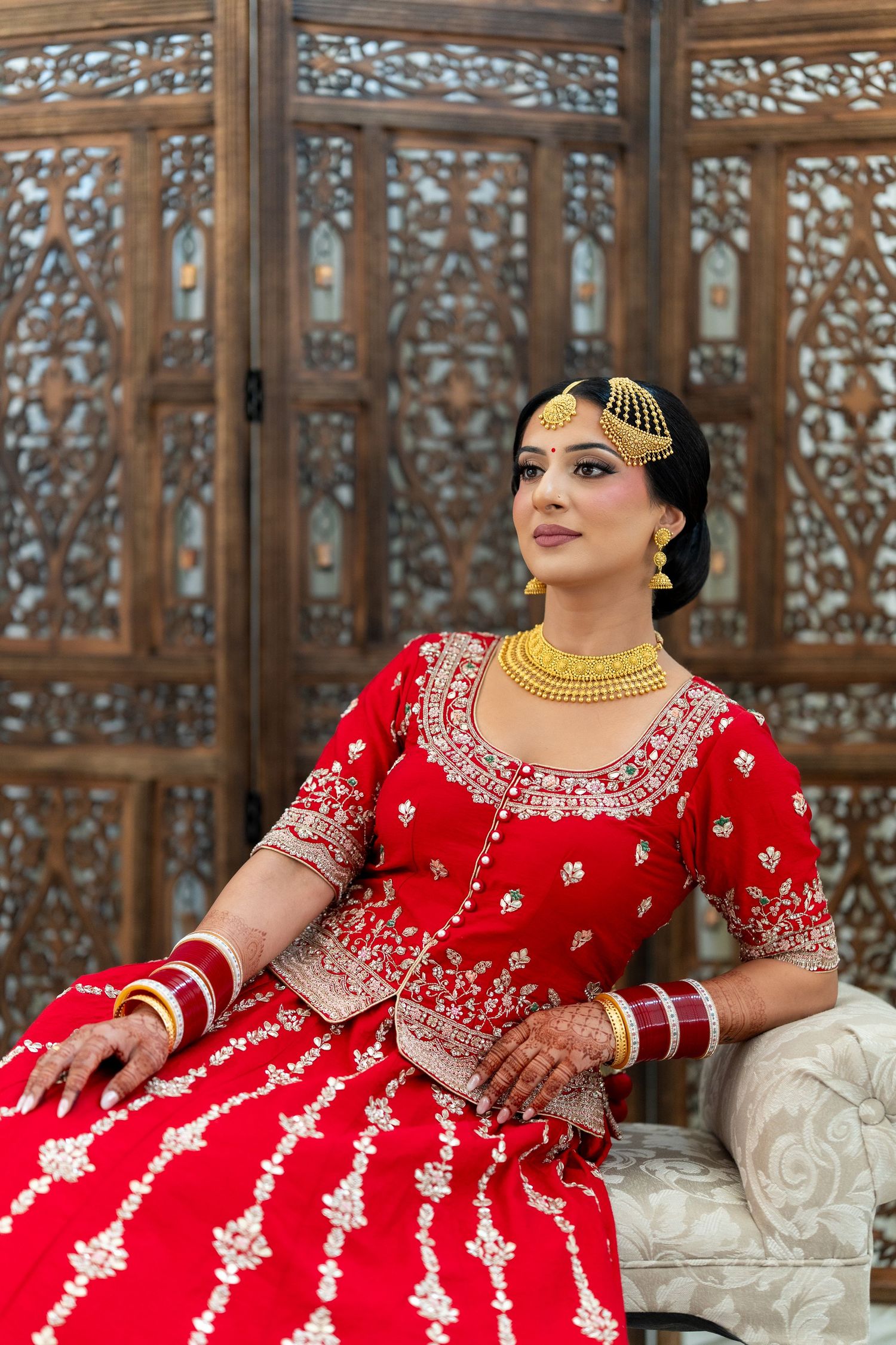 Traditional Indian bride in red embroidered lehenga poses elegantly against wooden carved backdrop.