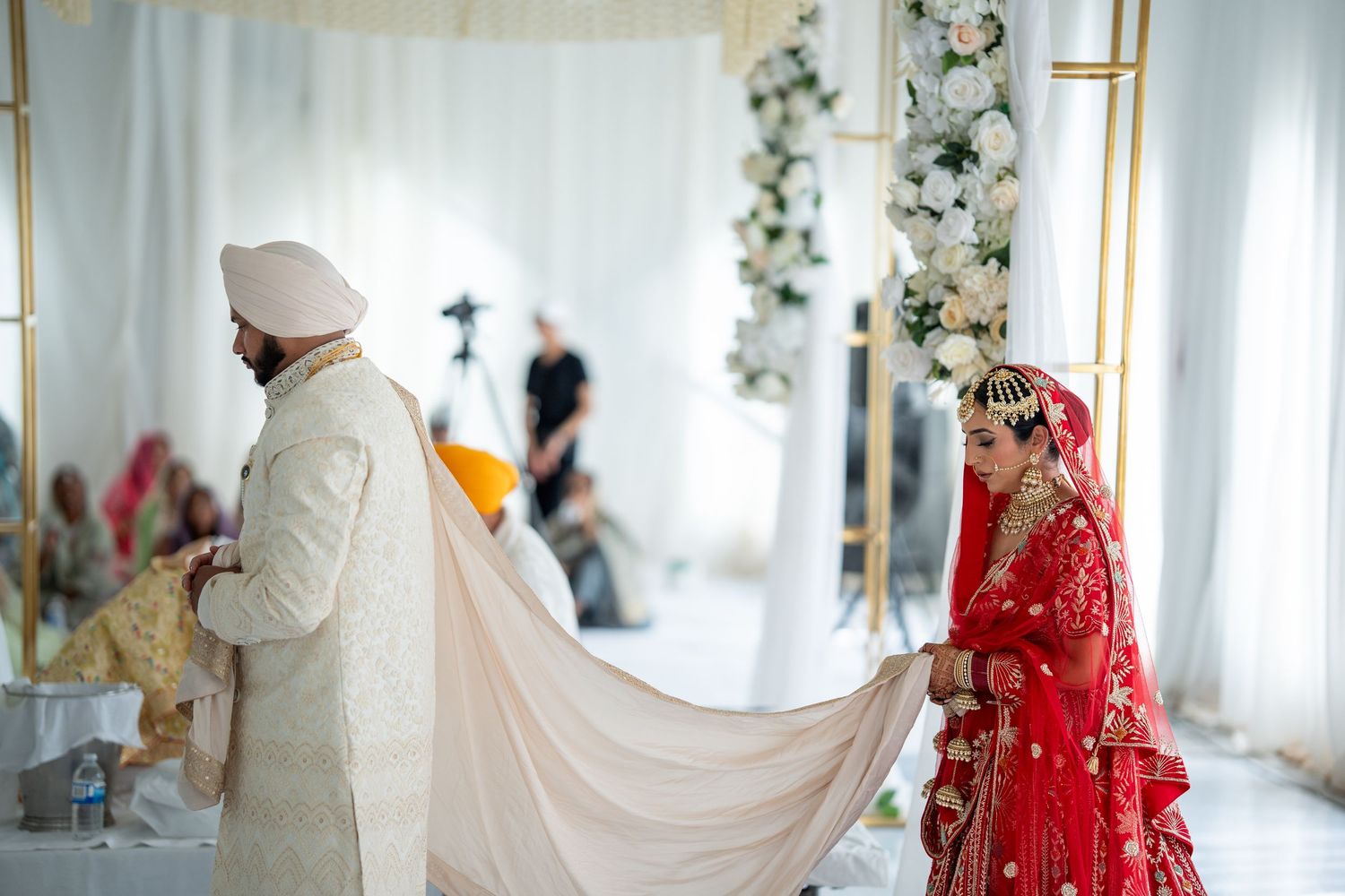 Wedding ceremony with couple in traditional Indian attire, bride in red and groom in white, under floral decor.