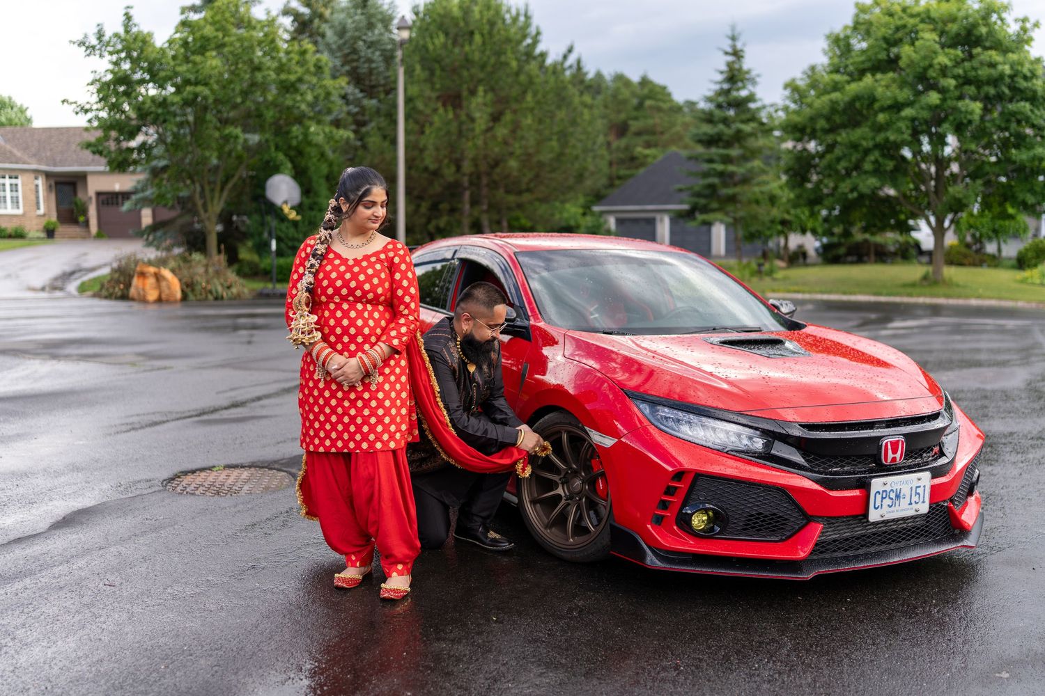 Woman in red salwar kameez stands beside a red Honda Civic Type R on a rainy suburban street.