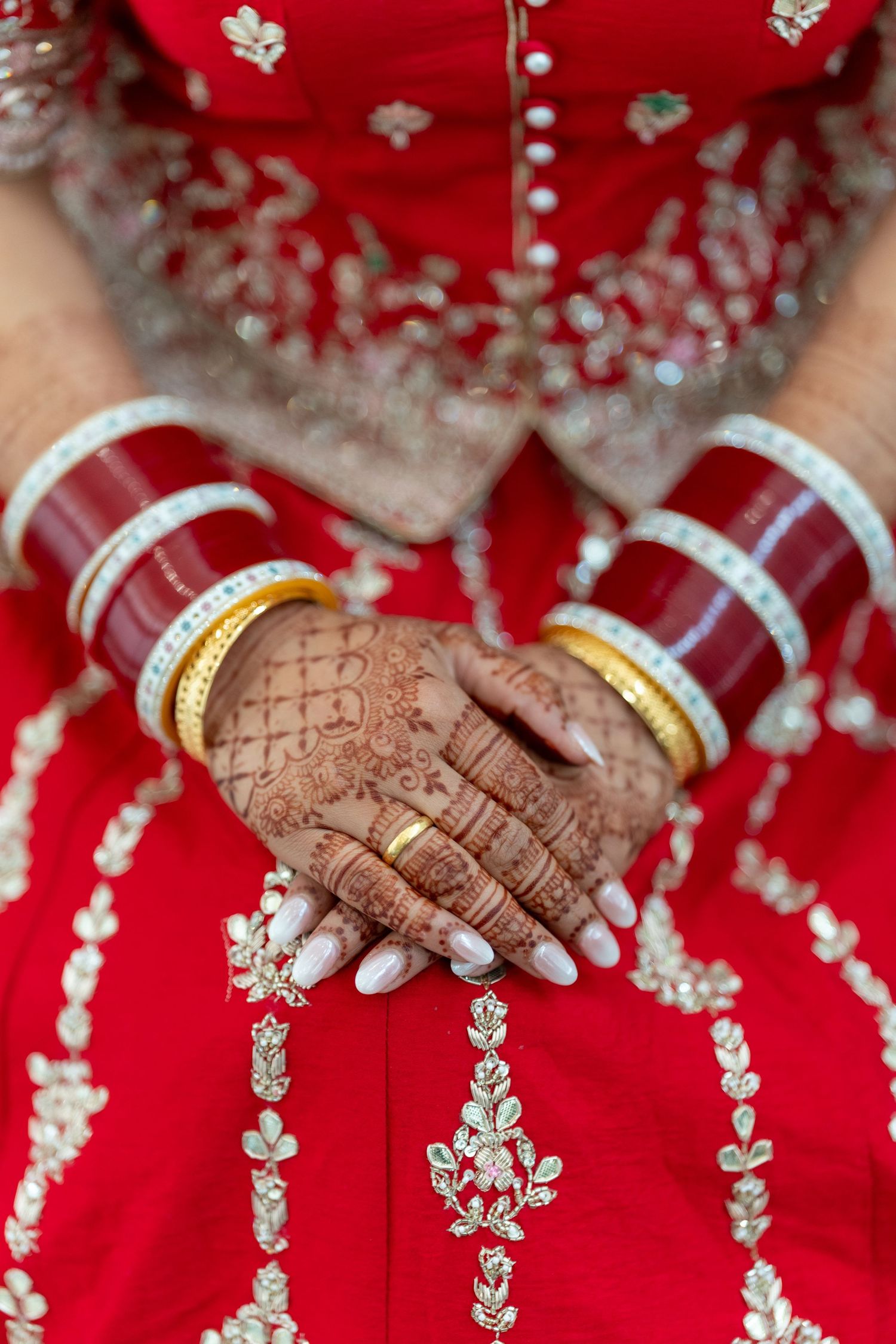 Close-up detail of traditional Indian bridal henna designs and jewelry on hands against a red background.