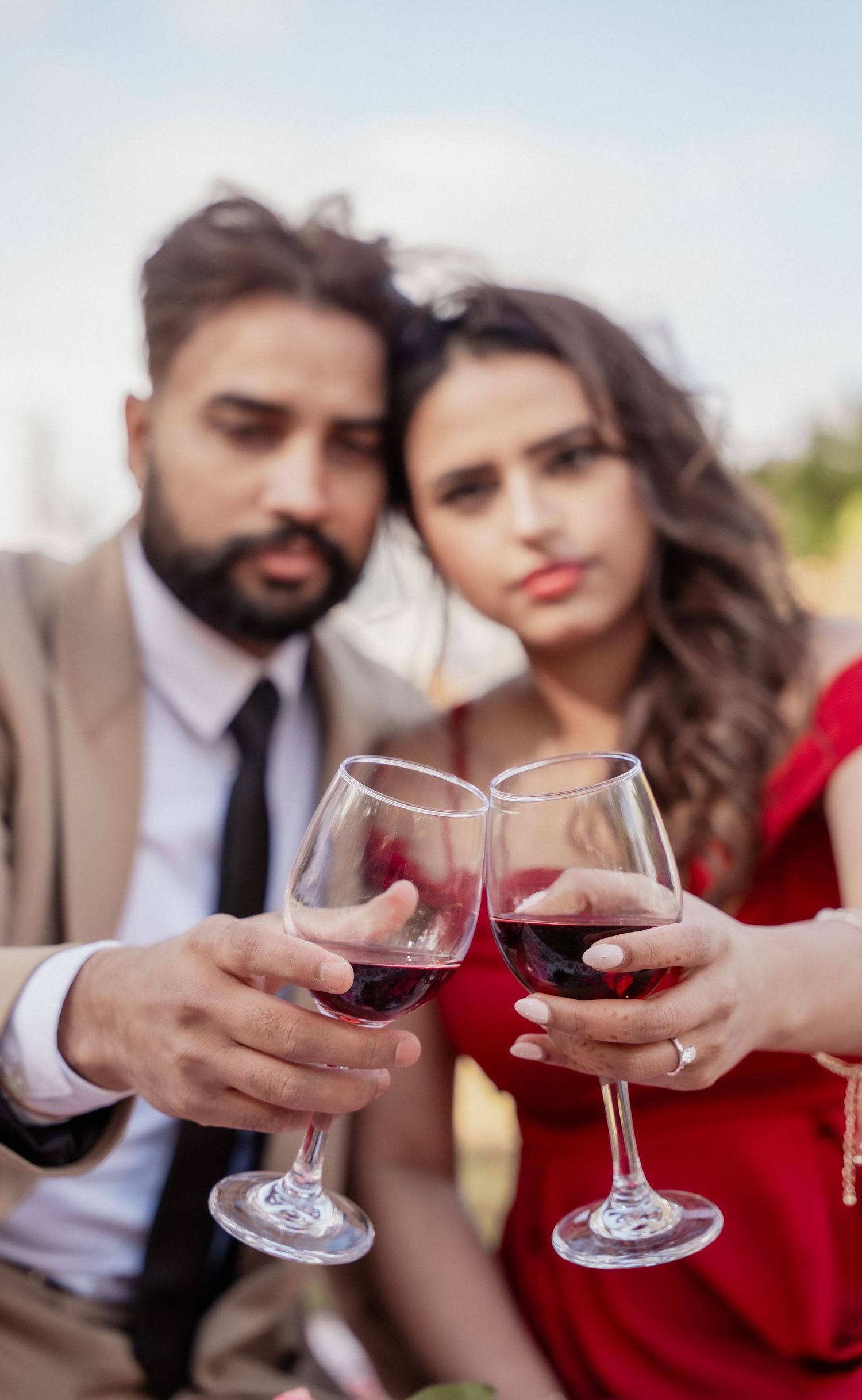 A couple in formal attire toasts with red wine glasses in a romantic outdoor setting.
