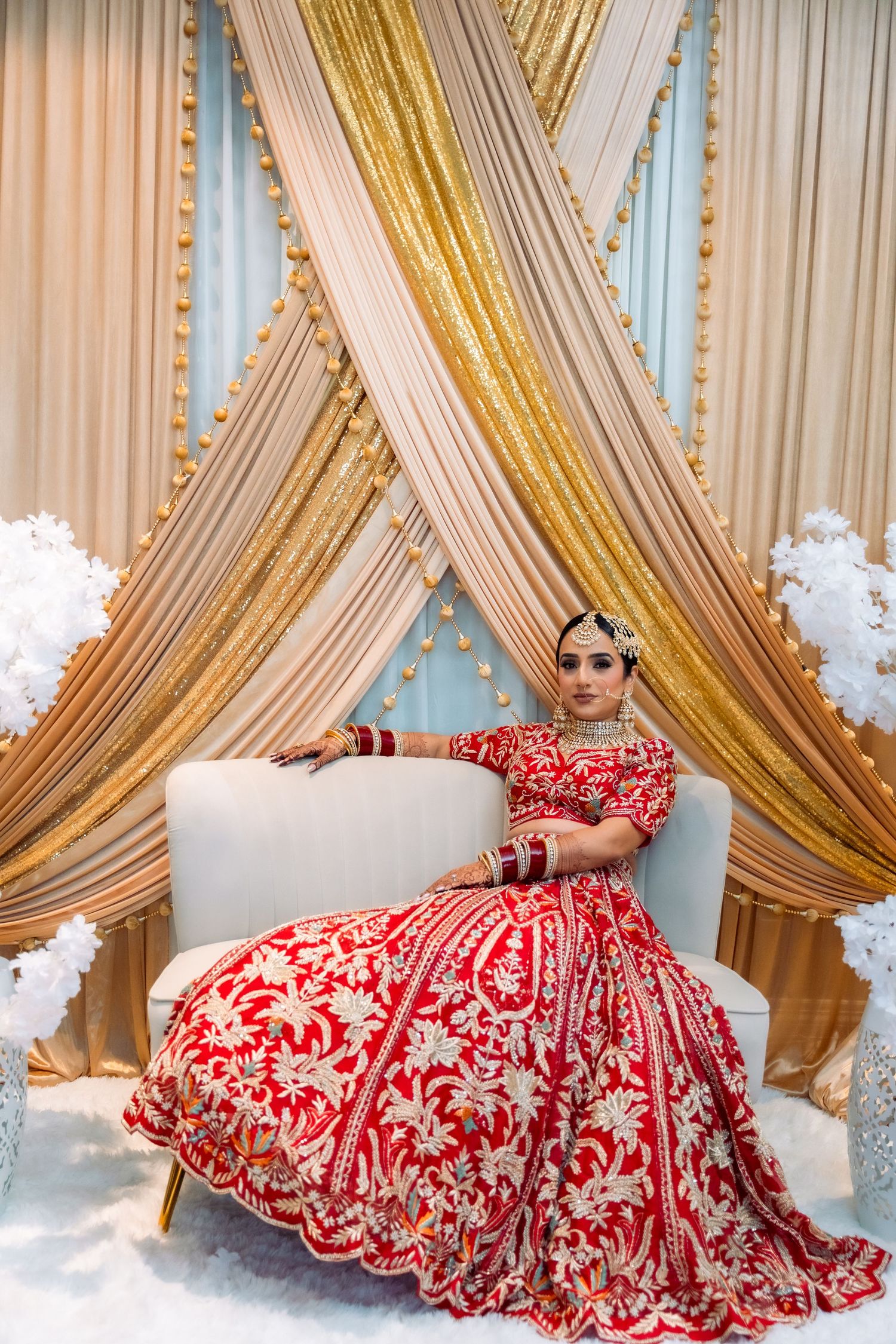 A red and white embroidered lehenga dress laid out on the floor with decorative gold drapes in the background.