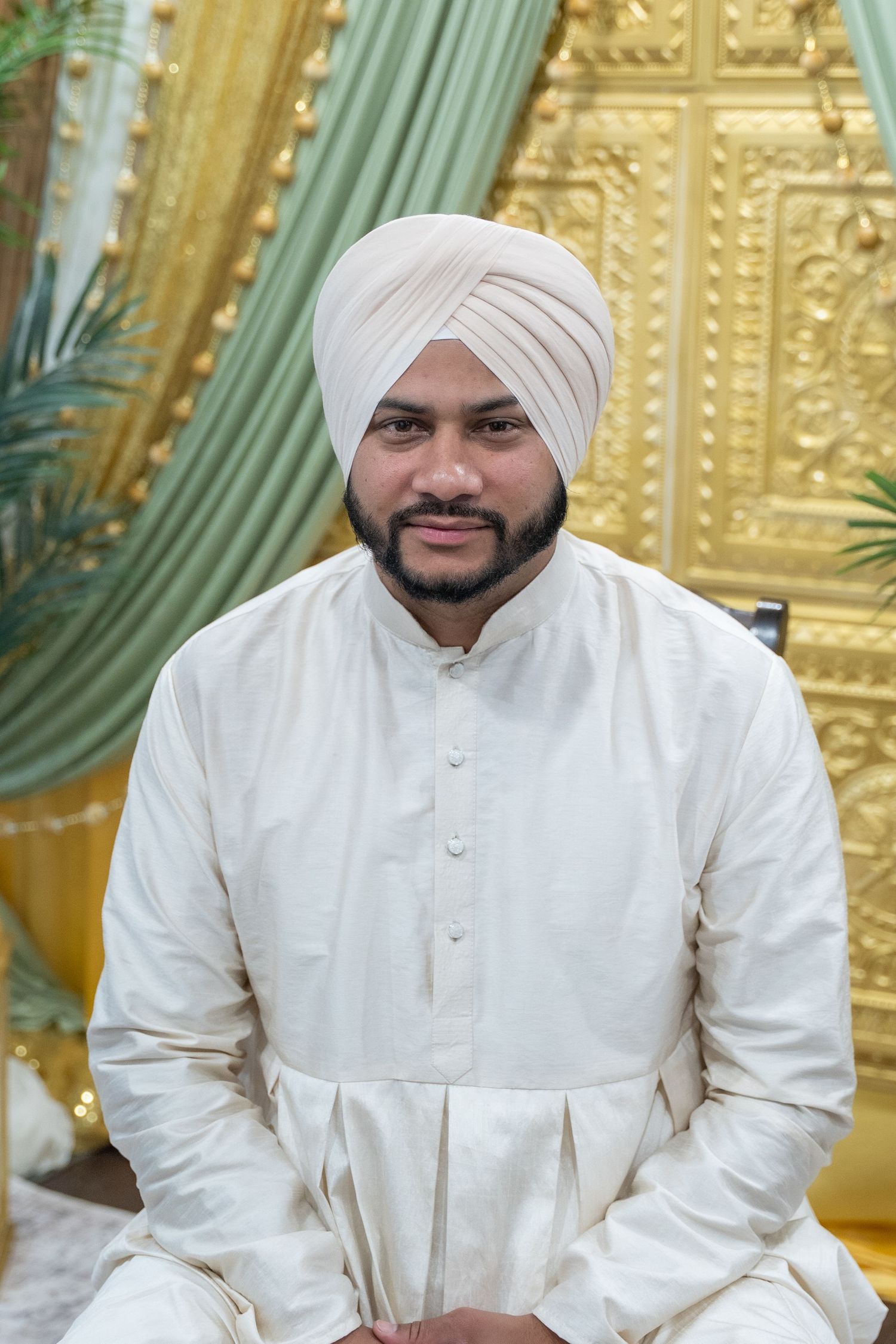 Person wearing traditional white Sikh wedding attire with turban against ornate gold backdrop.