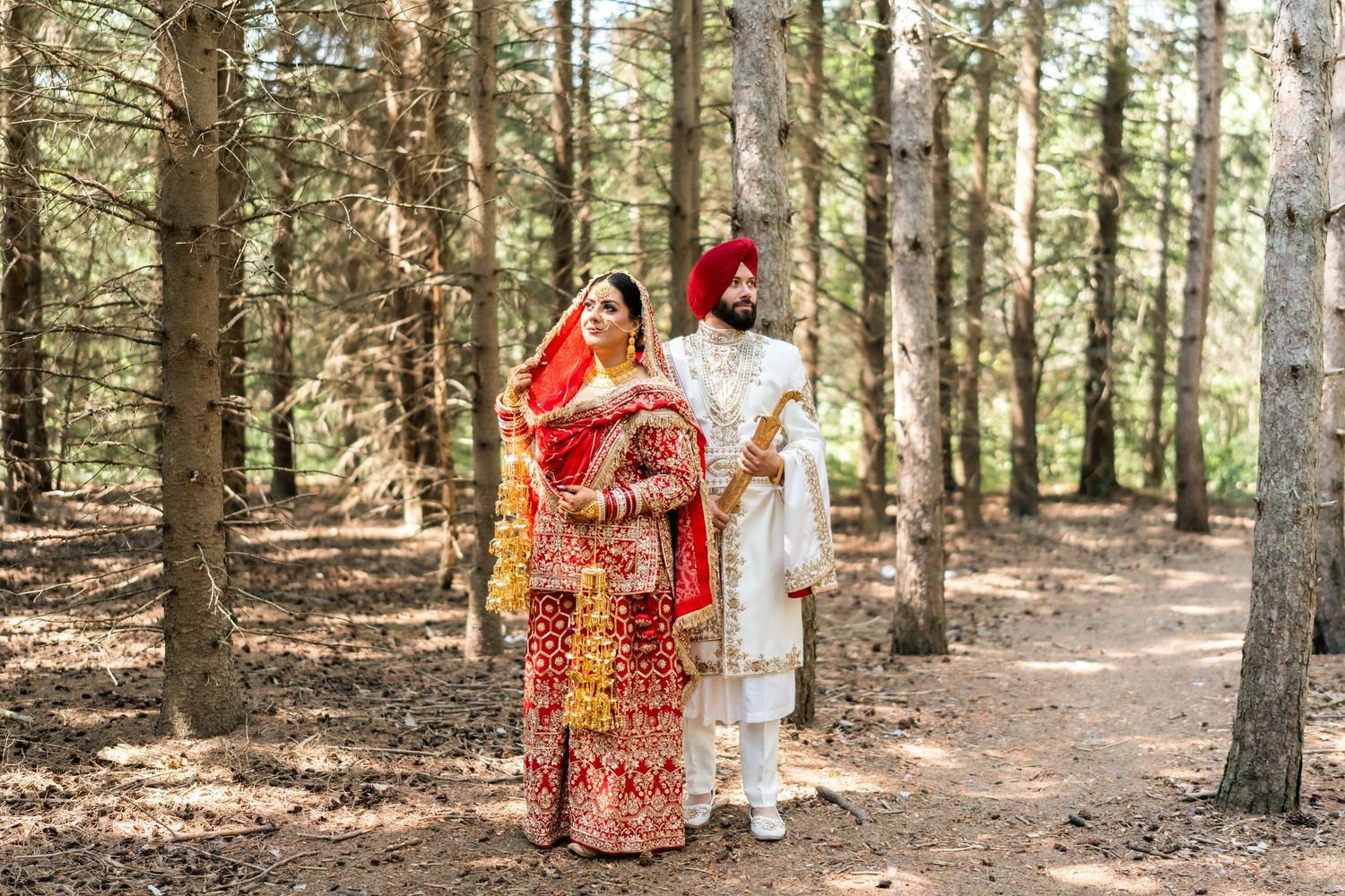 Sikh bride and groom in traditional red and white wedding attire standing among tall trees in a forest setting.