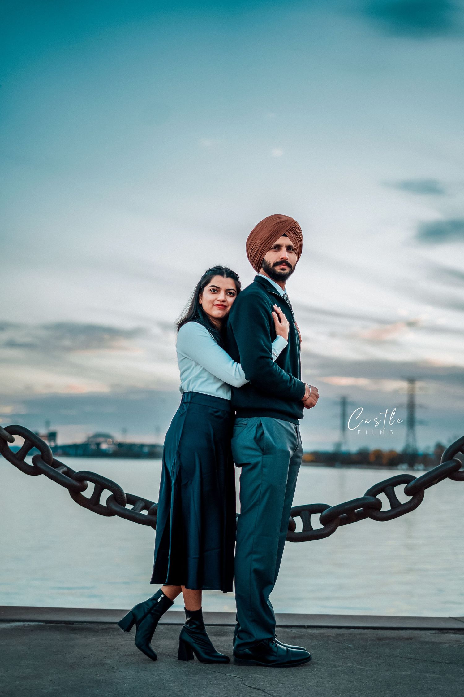 A couple poses for creative engagement photos with a large chain on a waterfront dock at sunset.
