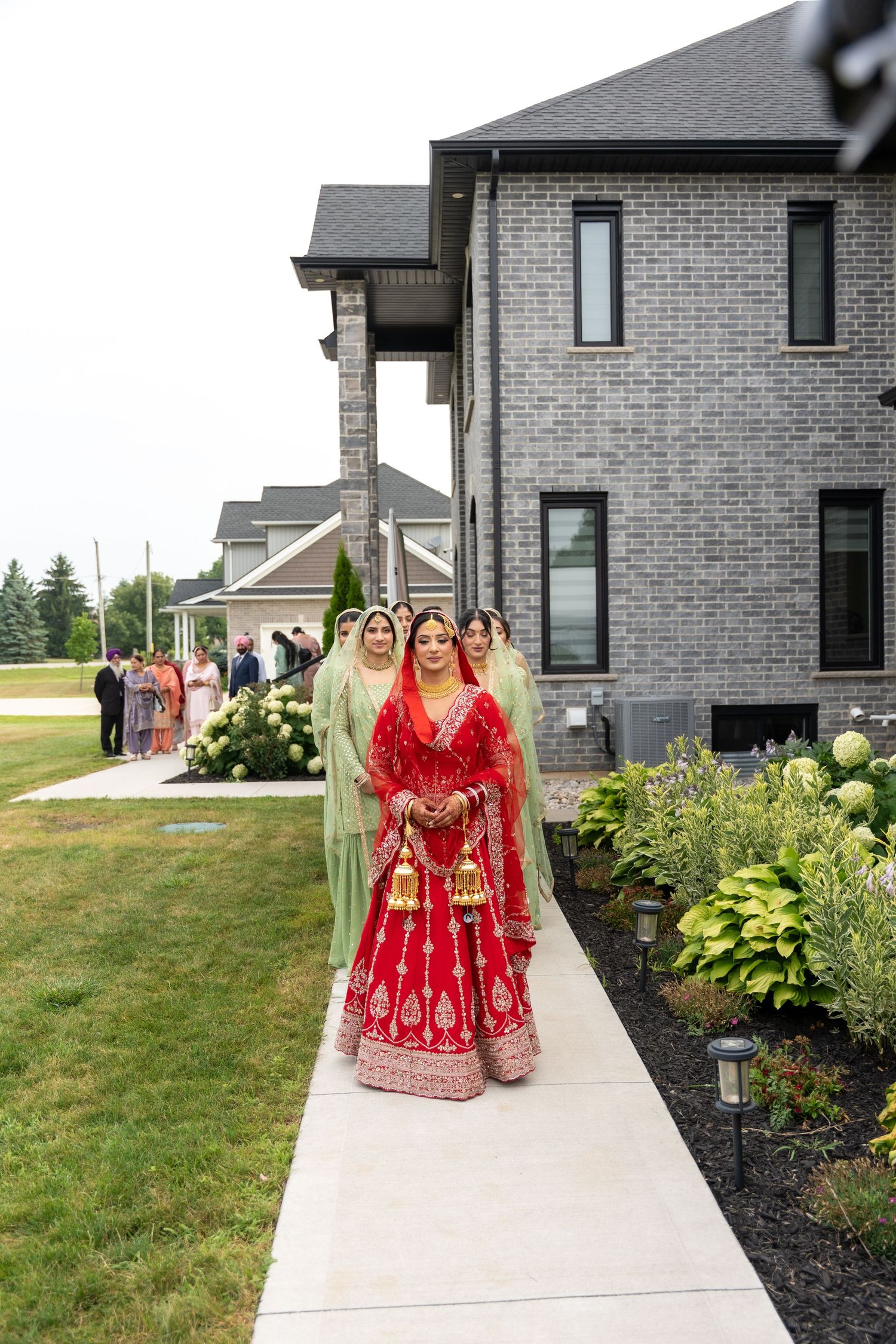 Red bridal lehenga photographed in sequence walking down a path outside a modern brick house.
