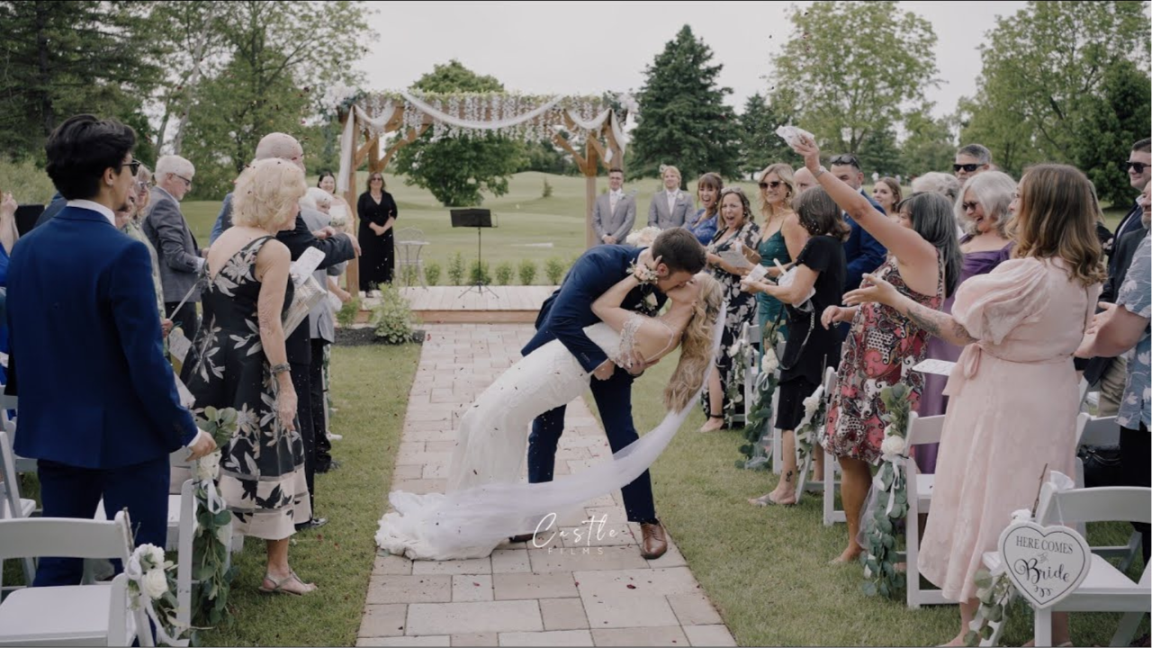 Bride and groom share romantic dip kiss while wedding guests celebrate with raised arms along outdoor ceremony aisle.