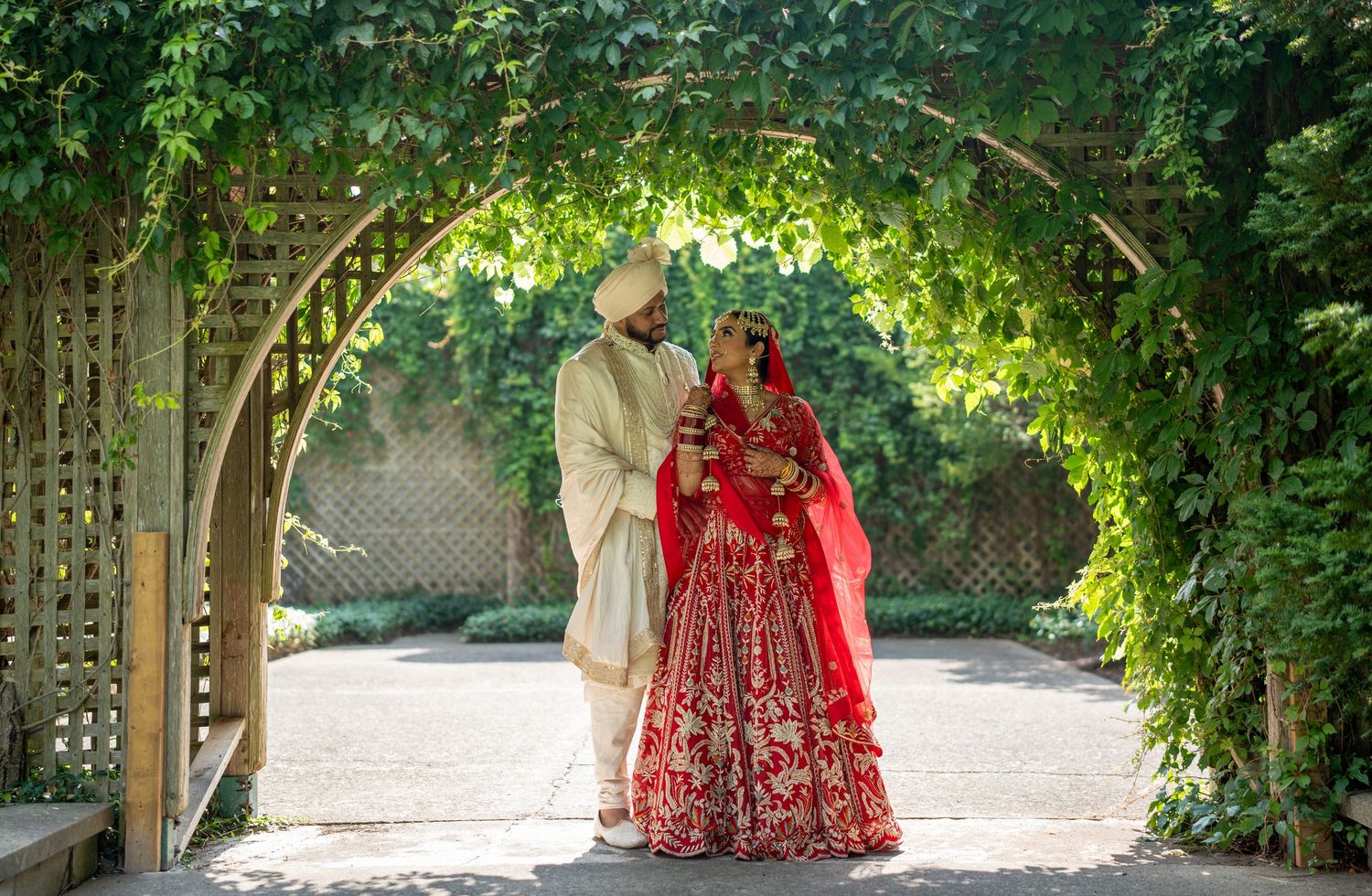 Traditional wedding couple stands beneath a lush green garden archway with the bride wearing an ornate red lehenga.