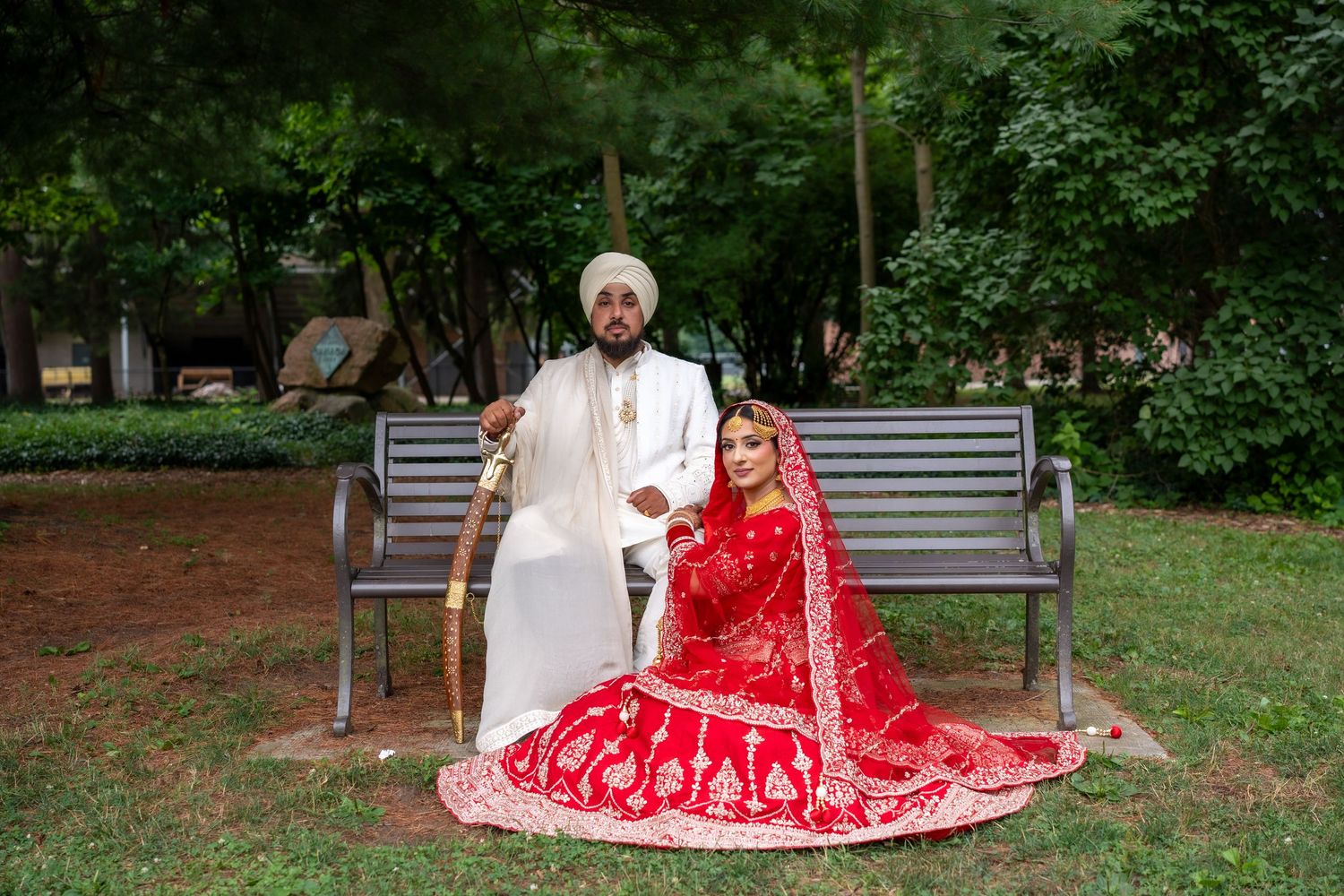 Traditional South Asian wedding couple sits together on park bench surrounded by lush greenery.