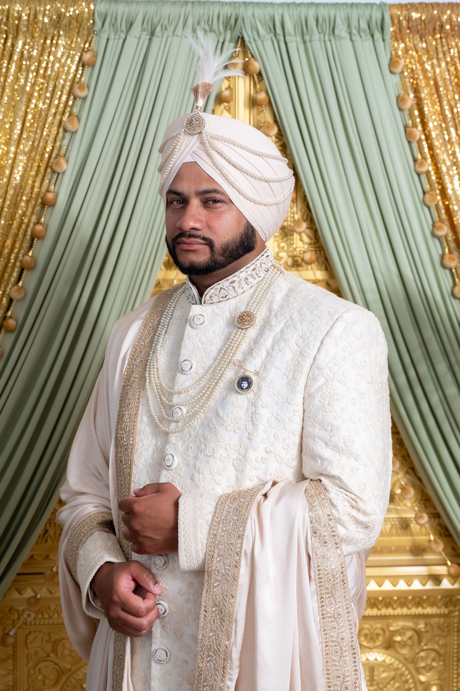 Groom in traditional white sherwani with gold embroidery and turban poses against gold and mint backdrop.