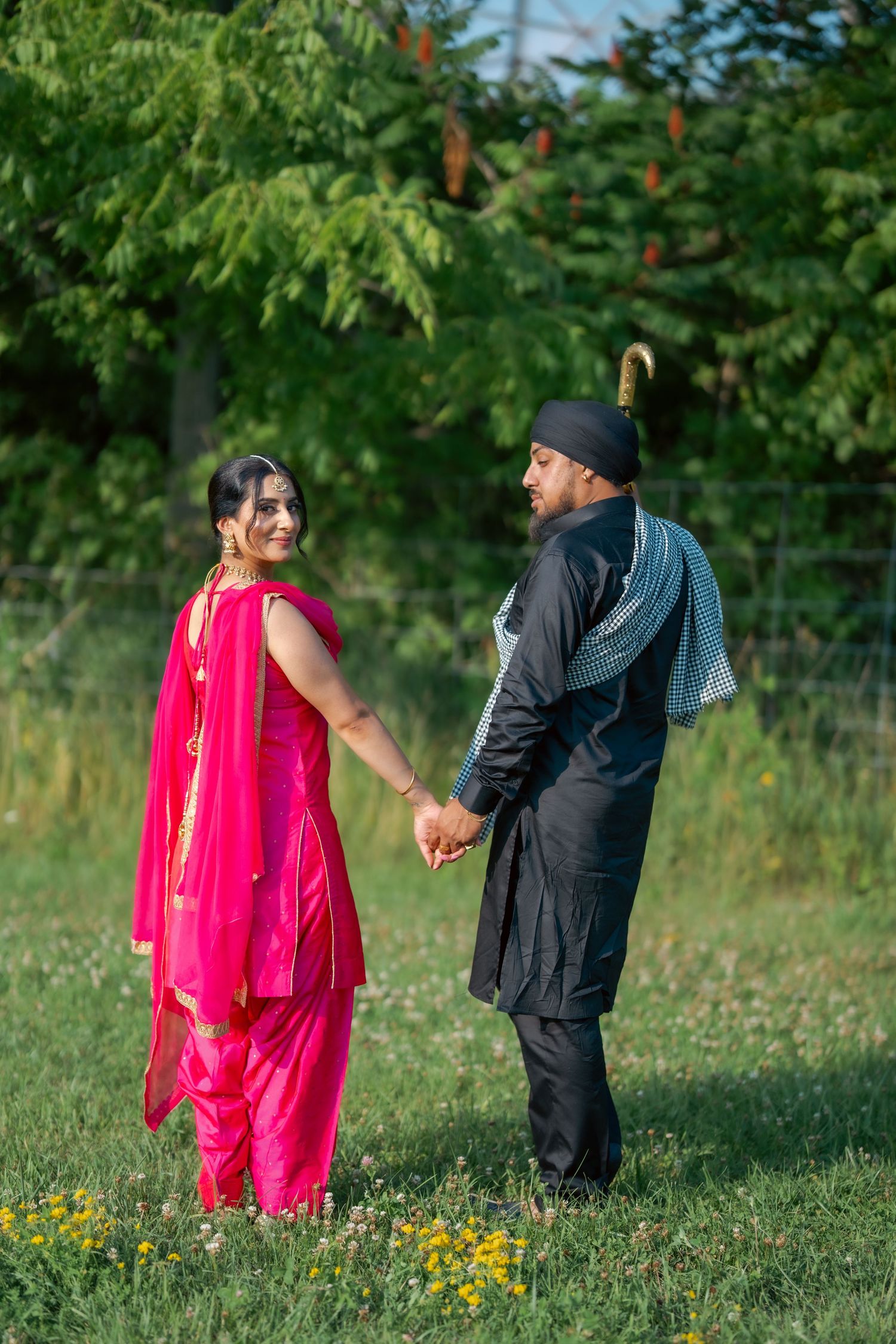 A couple takes romantic photos in a lush green garden setting with bright pink traditional outfits.