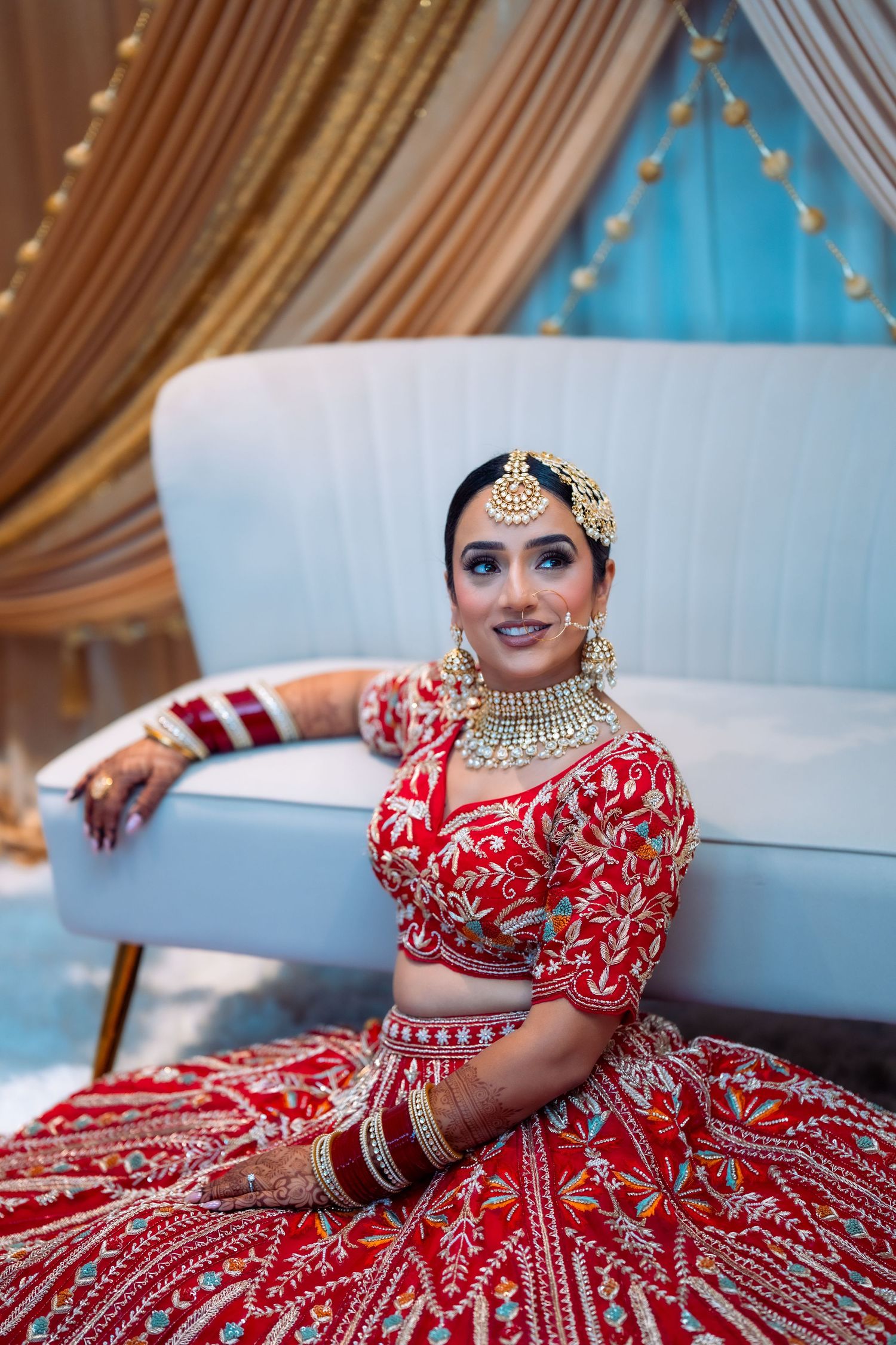 Bride wearing ornate red and gold lehenga with traditional Indian bridal jewelry sits gracefully.