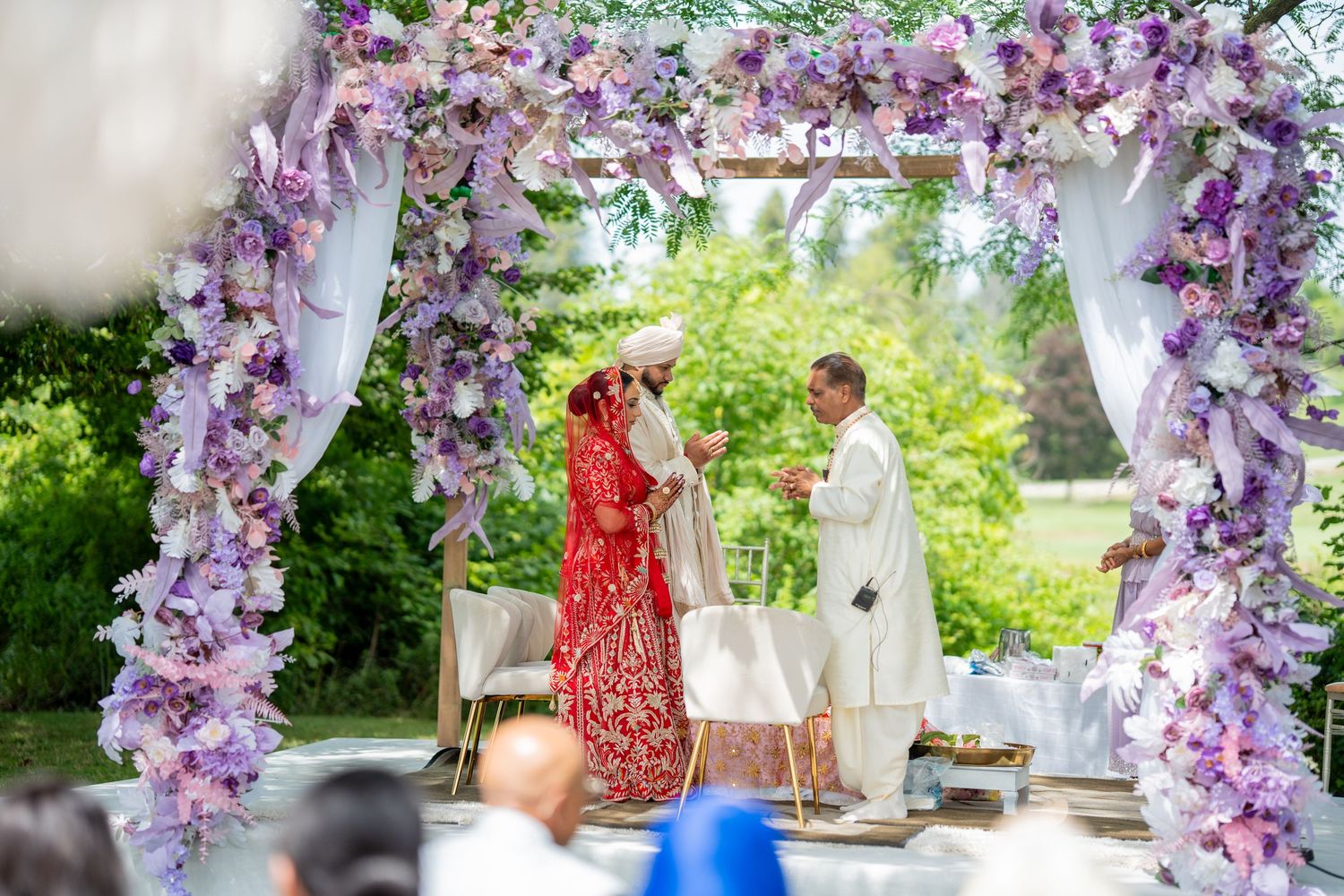 Outdoor wedding ceremony setup with purple floral arch and white decor elements.