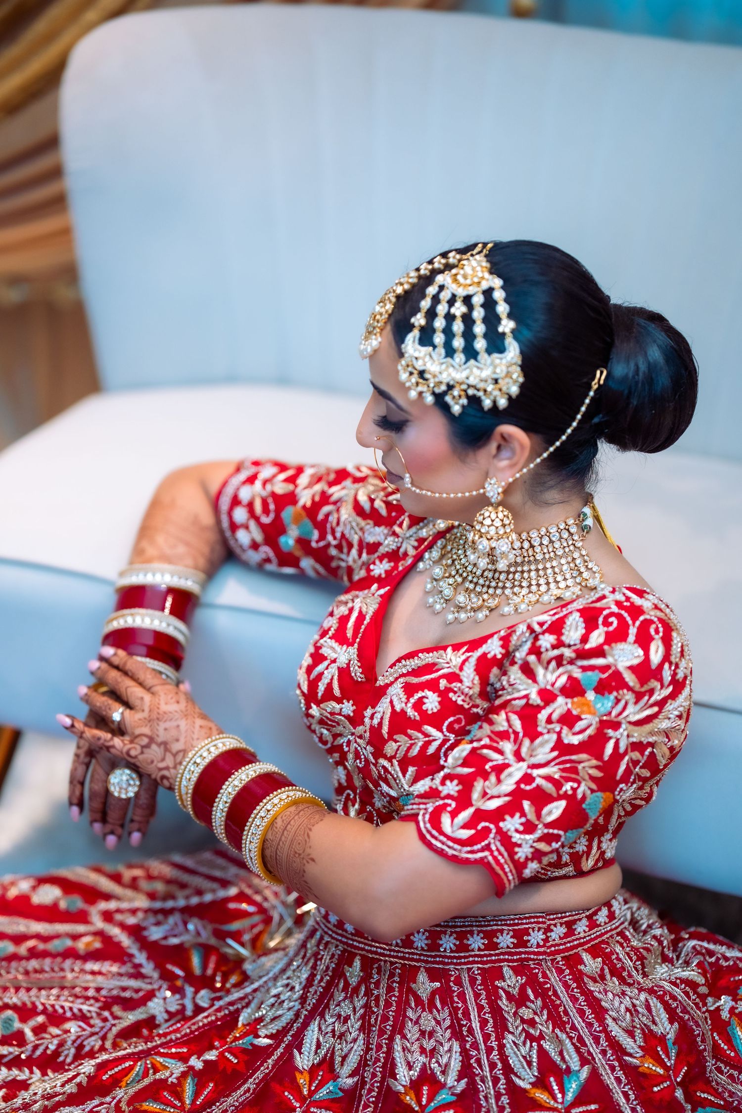 Traditional Indian bride in red lehenga with intricate embroidery and gold jewelry poses elegantly on a couch.