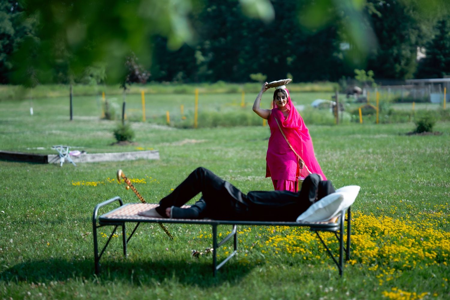 Person in bright pink shirt relaxes on outdoor cot surrounded by yellow wildflowers in grassy park setting.