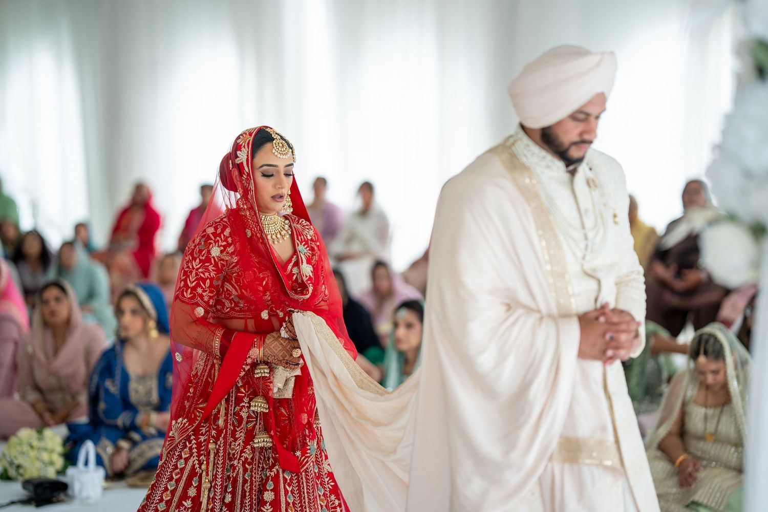 Traditional Indian wedding ceremony with bride in red lehenga and groom in white sherwani under white tent.