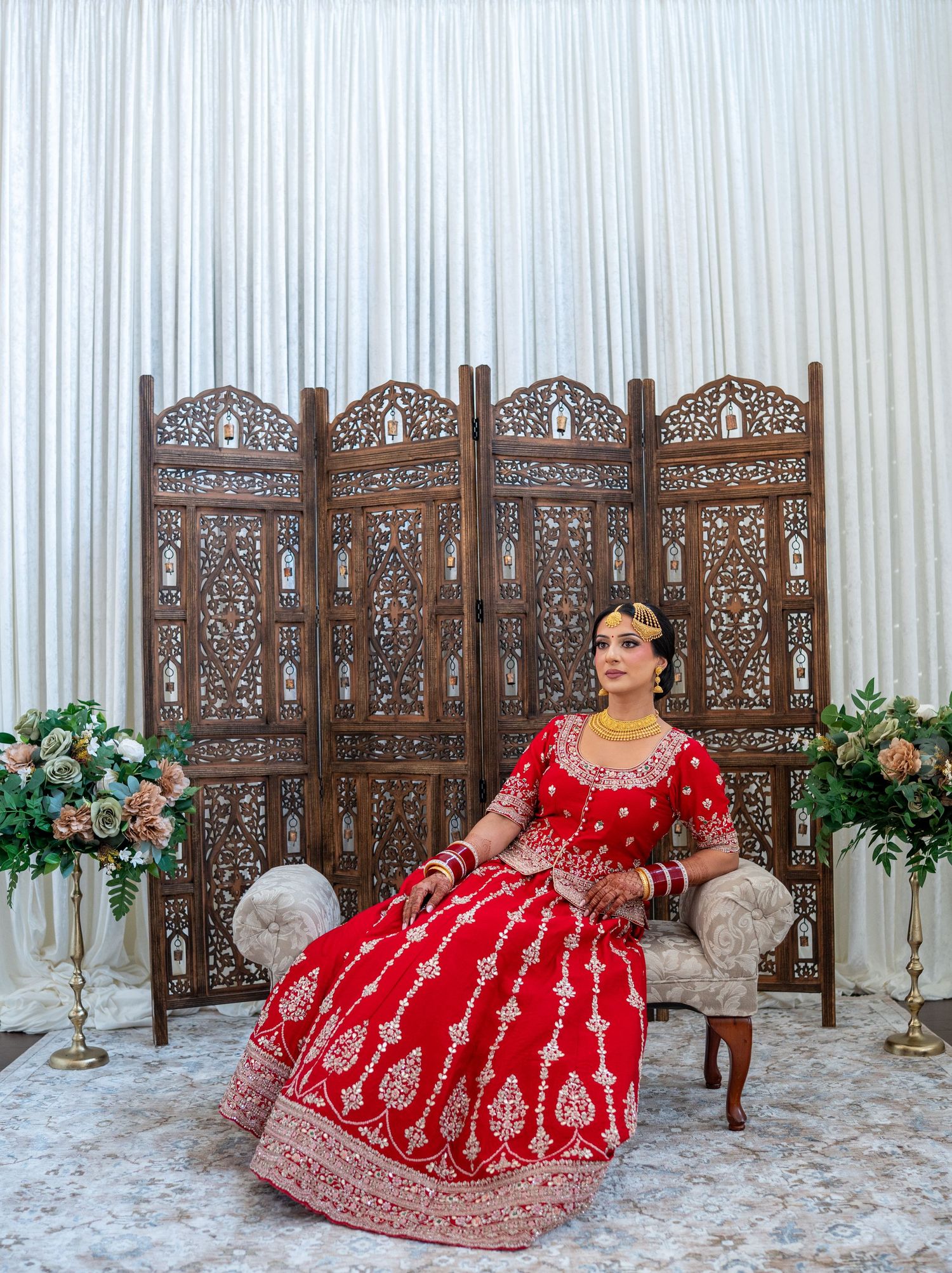 Traditional Indian bridal attire featuring a red embroidered lehenga against an ornate wooden screen with floral arrangements.