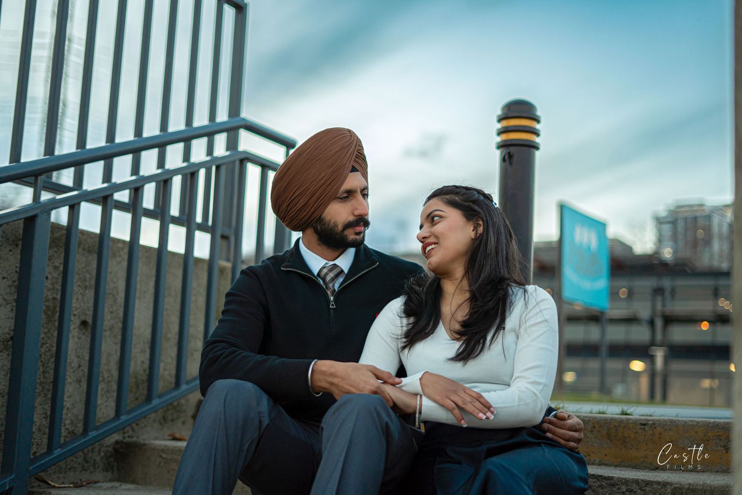 A couple shares a romantic moment while sitting on a metal railing against an urban backdrop at dusk.