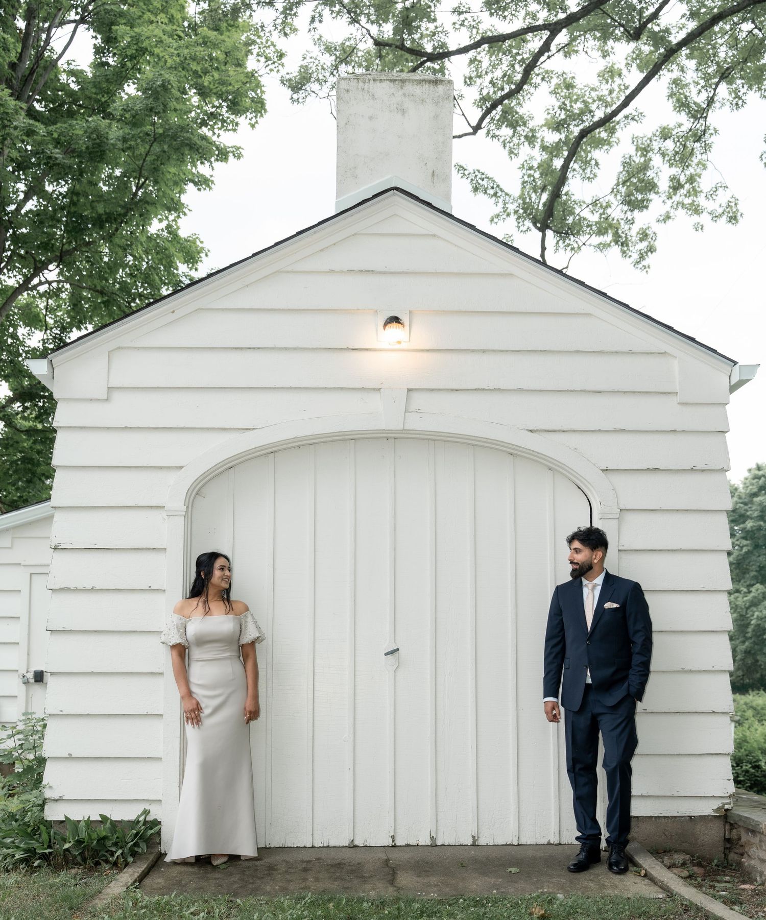 A couple poses on either side of a white arched barn door entrance with a glowing light fixture above.