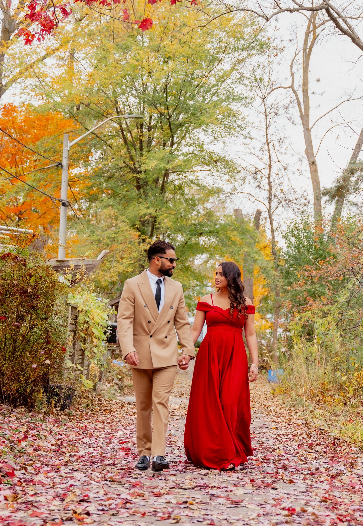 A couple in formal attire walks through vibrant autumn foliage with red and yellow leaves scattered on the ground.
