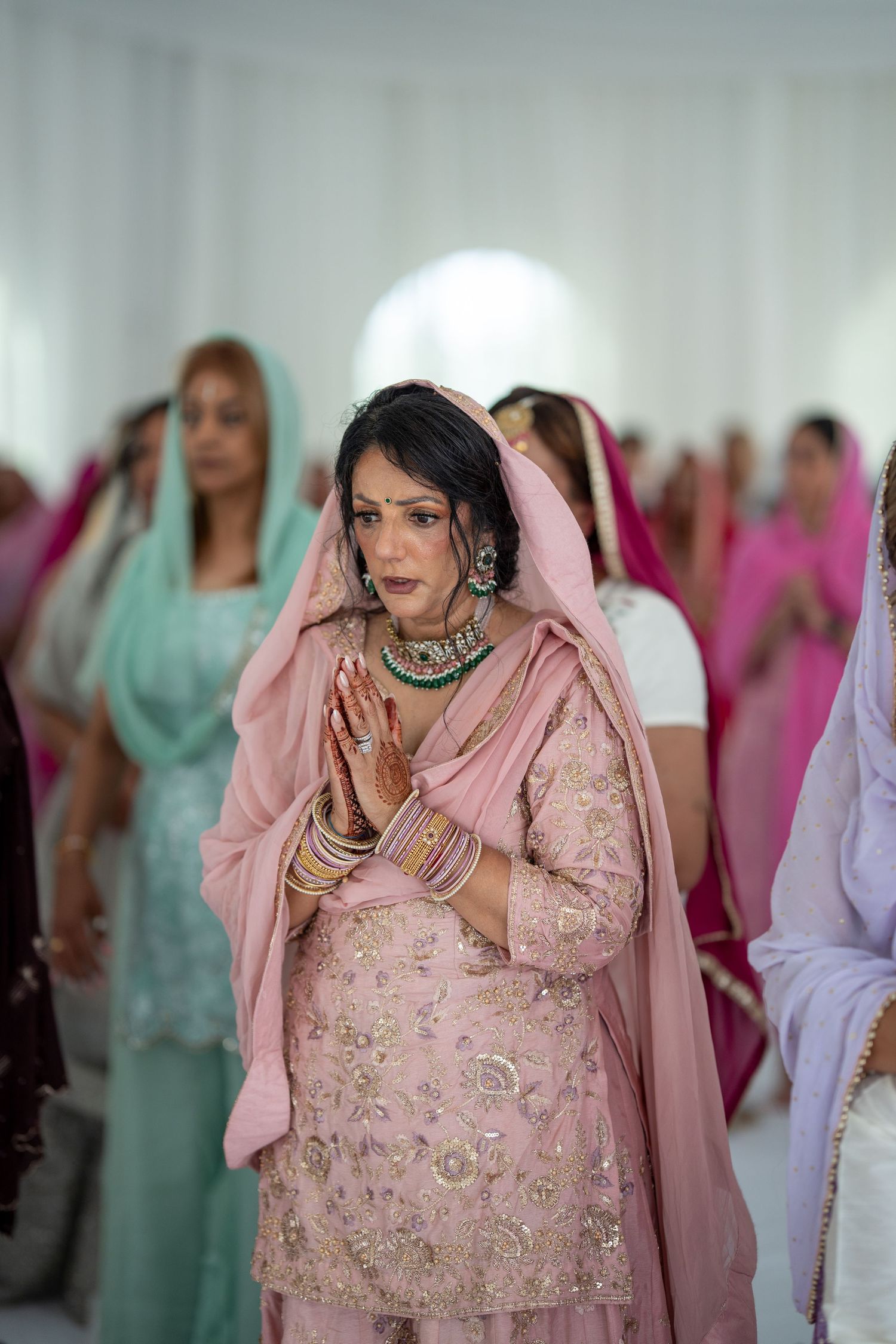Person in pink embroidered traditional dress performing prayer gestures during ceremony.