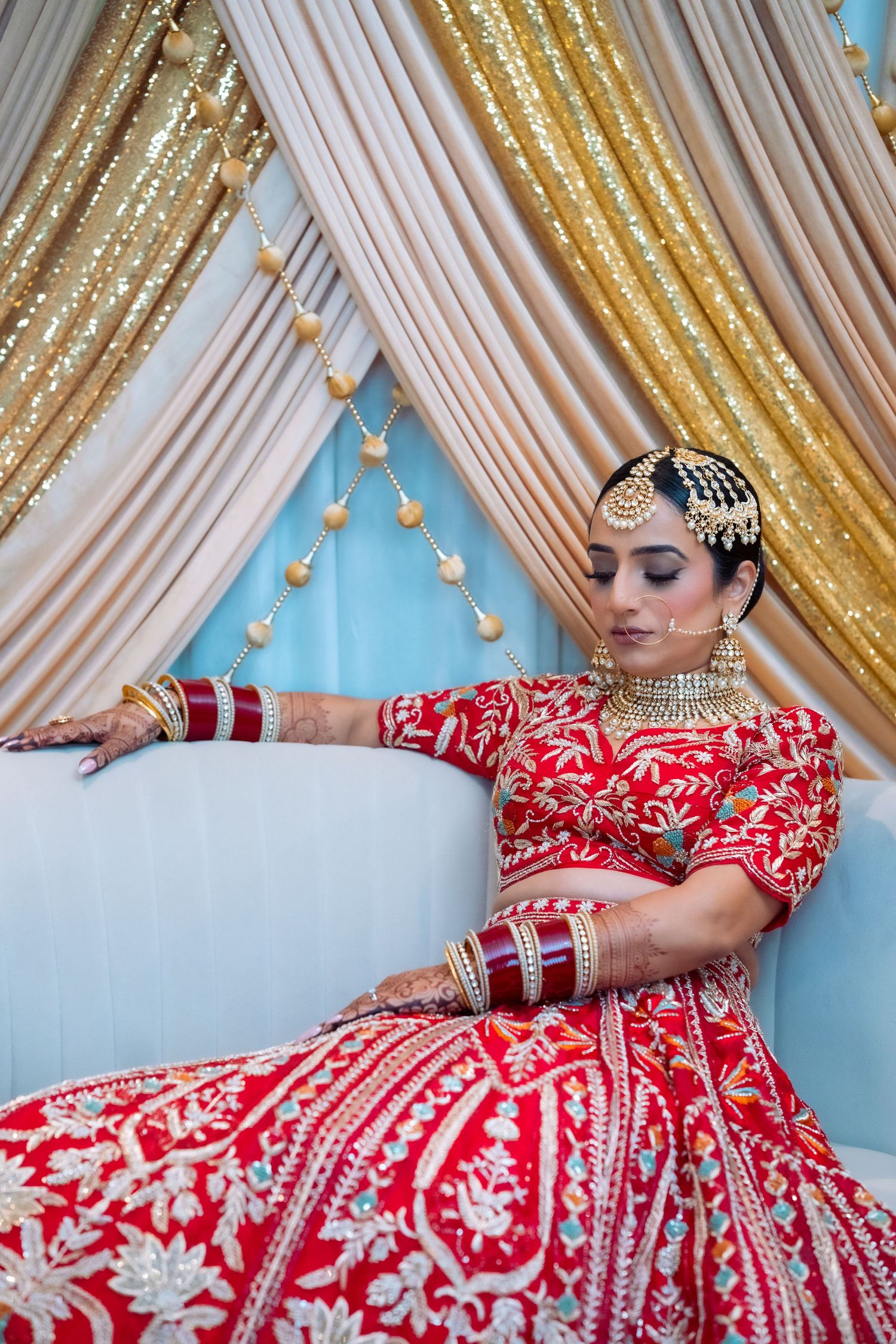 Traditional South Asian bride in red and white embroidered lehenga poses elegantly against gold and blue backdrop.