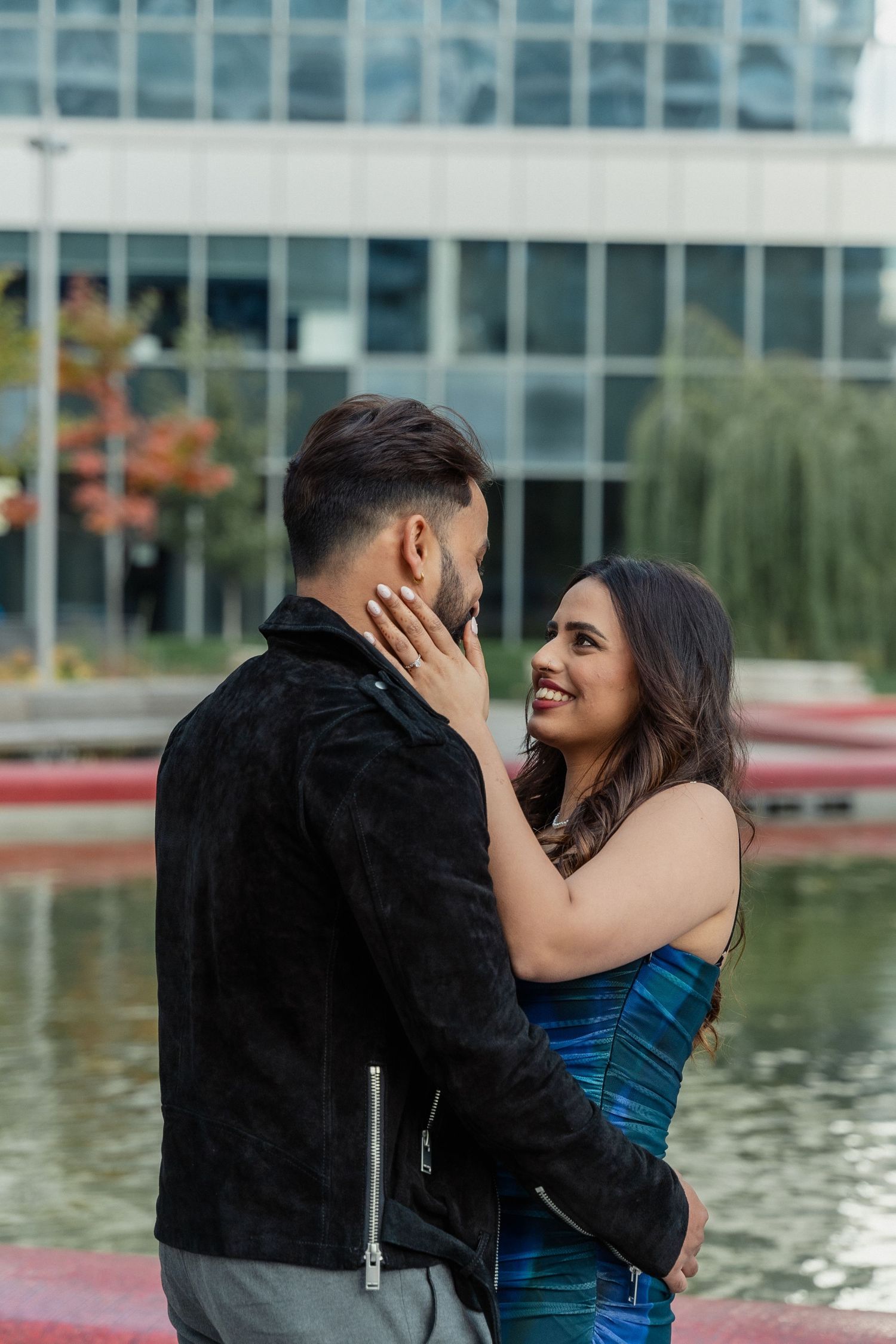 Two people embrace by a waterfront with modern glass buildings in the background.