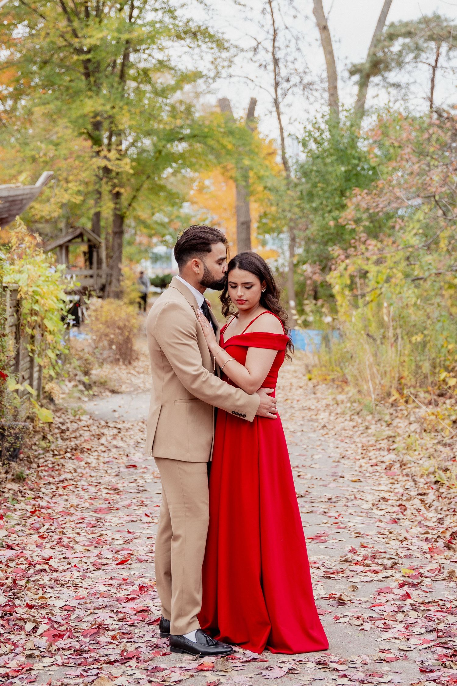 A couple in formal attire shares romantic moments on a leaf-covered autumn path surrounded by fall foliage.
