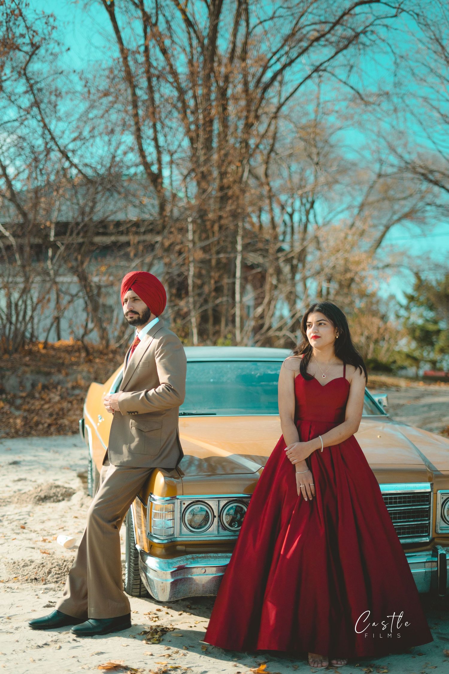 A couple poses together near a vintage turquoise car, with the woman wearing a red dress and the man in brown attire.