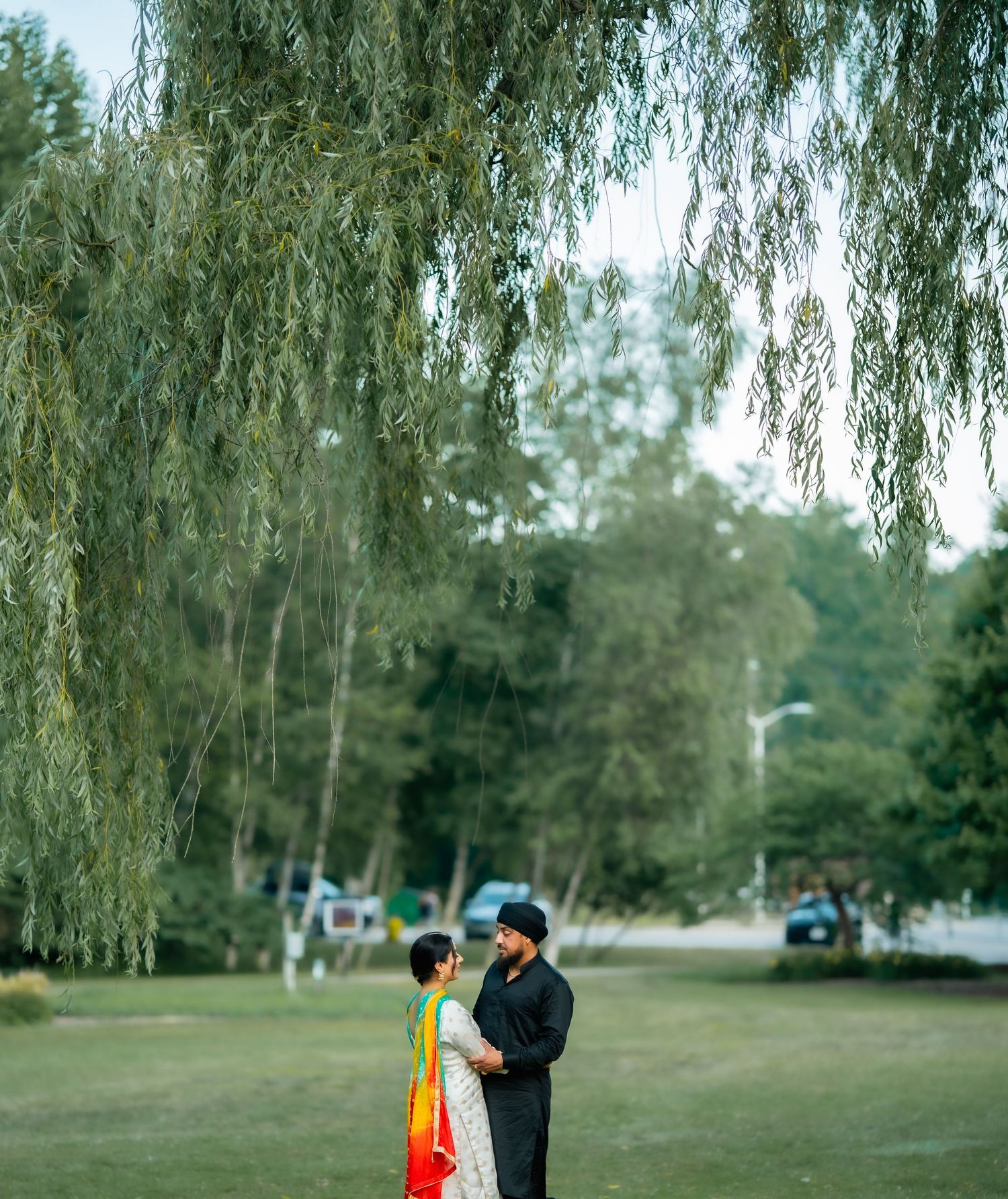 Two people stand together under drooping willow trees in a serene park setting.