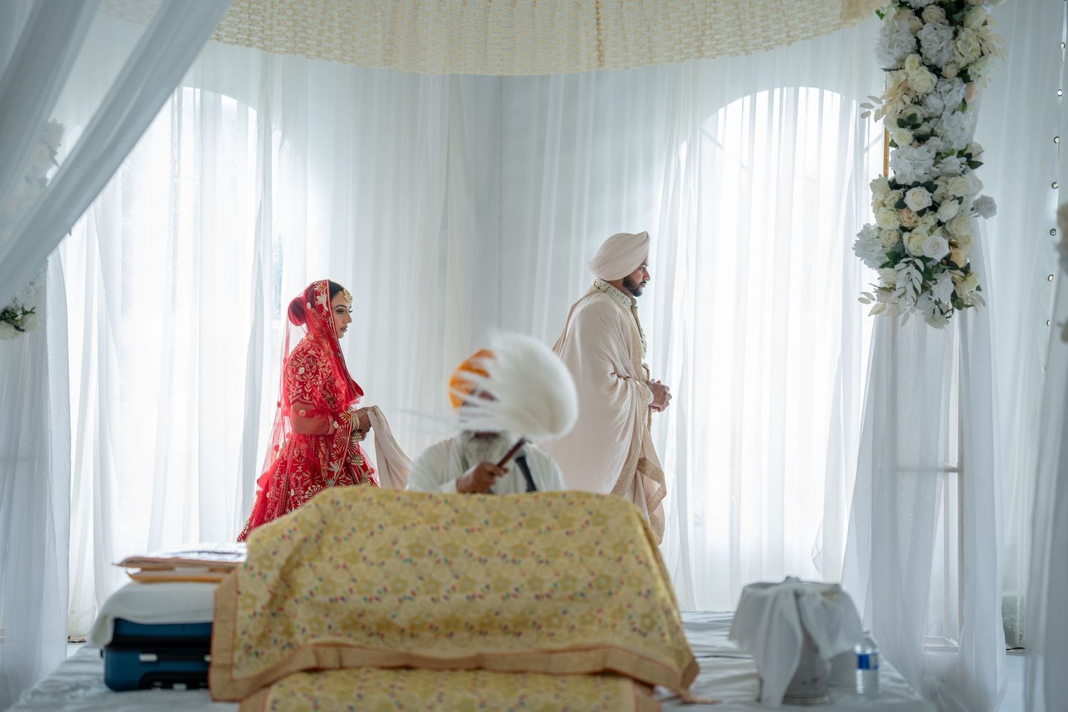 Sikh wedding ceremony with bride in red lehenga and groom in white turban standing near decorated mandap.
