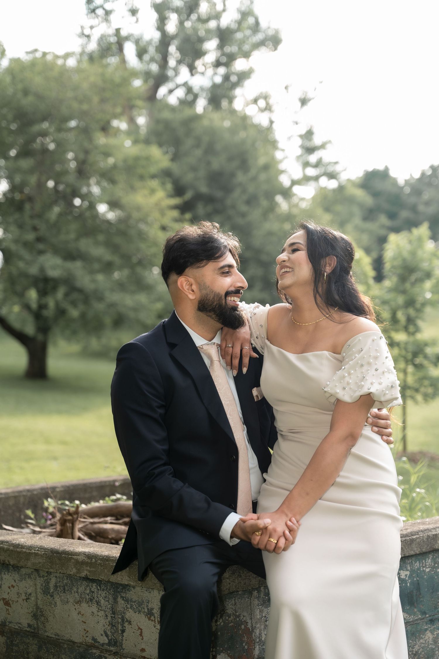 Newlywed couple shares joyful laughter together while posing for romantic outdoor wedding portraits in a garden setting.