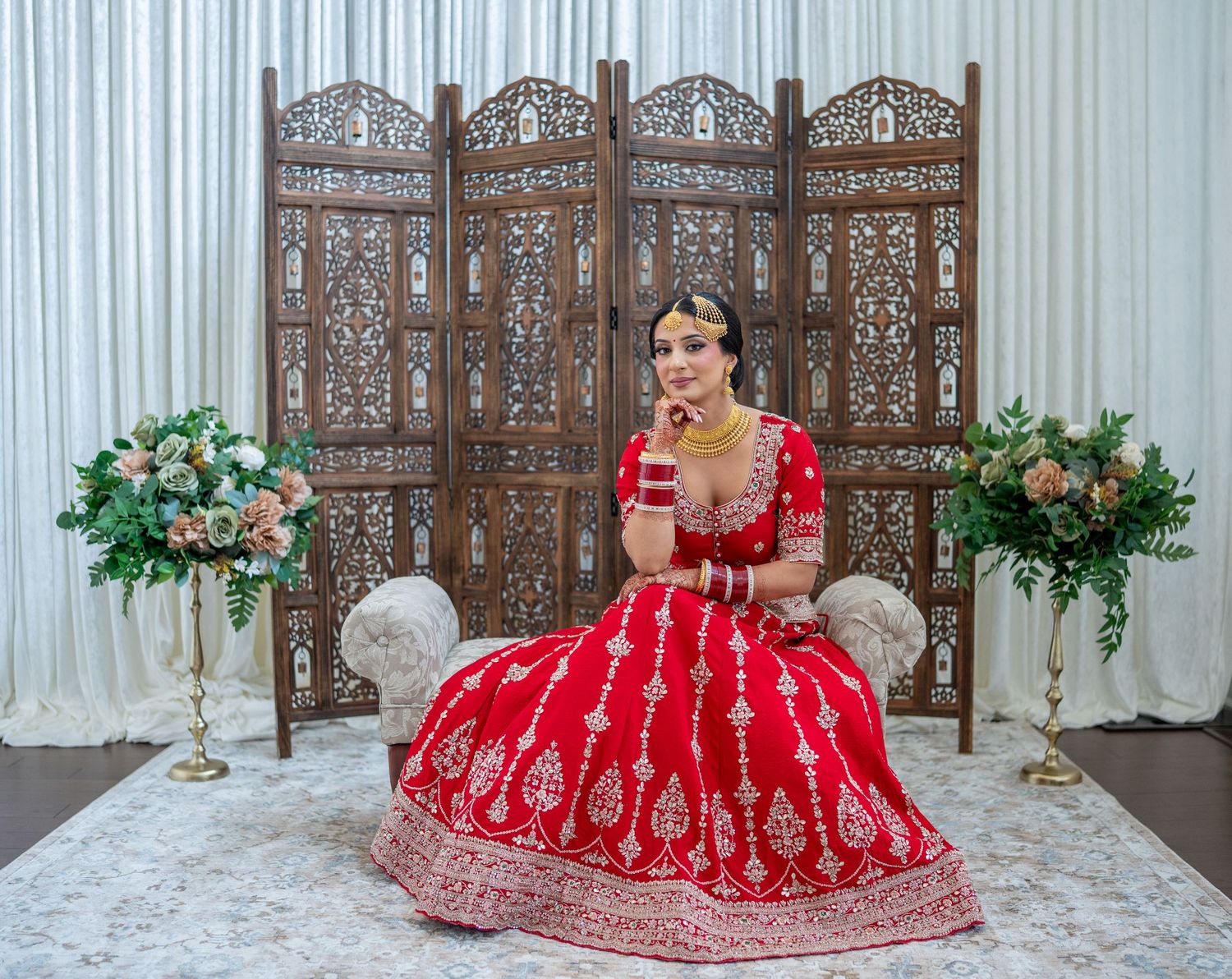 A traditional Indian bride in a stunning red lehenga poses elegantly against an ornate wooden screen backdrop.