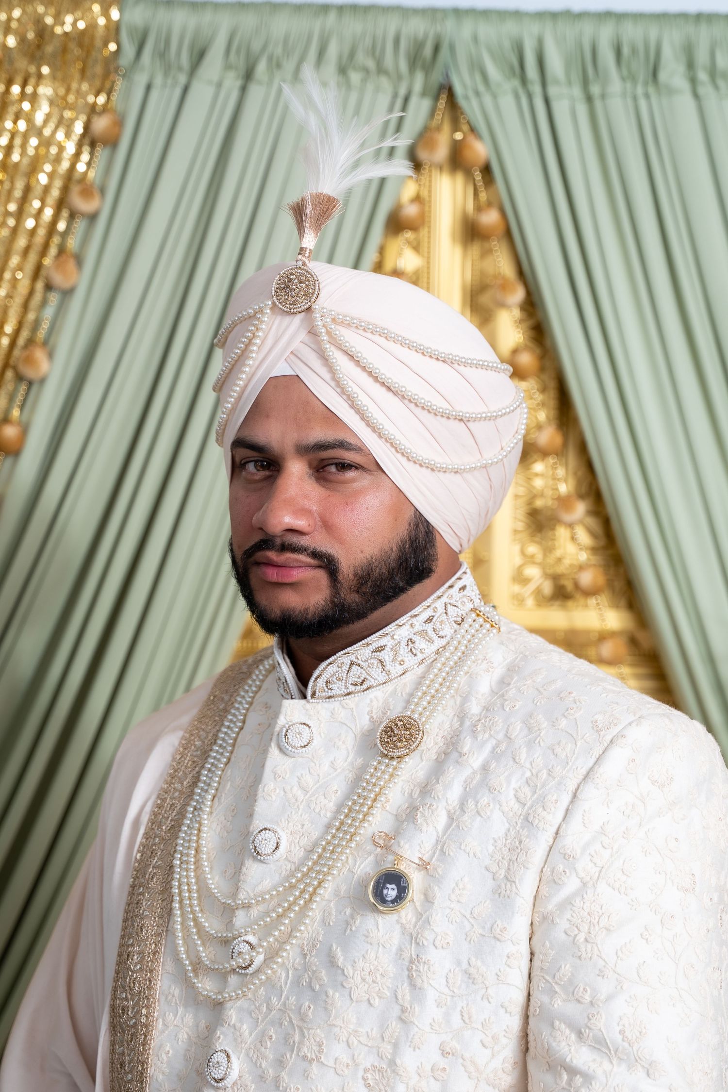 Traditional Indian groom wearing an ornate white sherwani and turban with pearl necklaces against mint green backdrop.