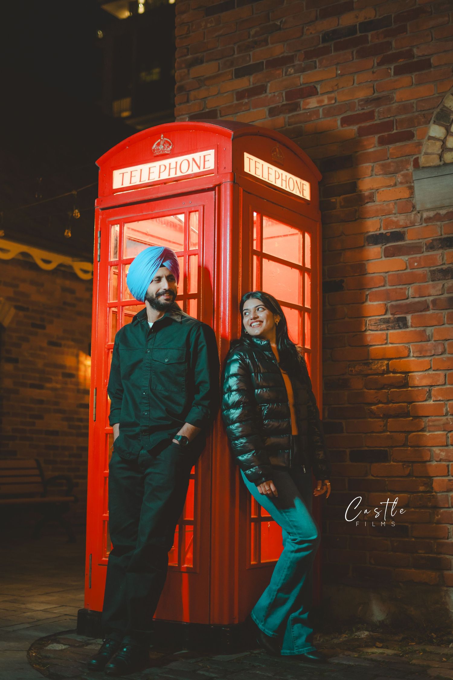 Two people pose casually next to an iconic red British telephone booth against a brick wall at night.