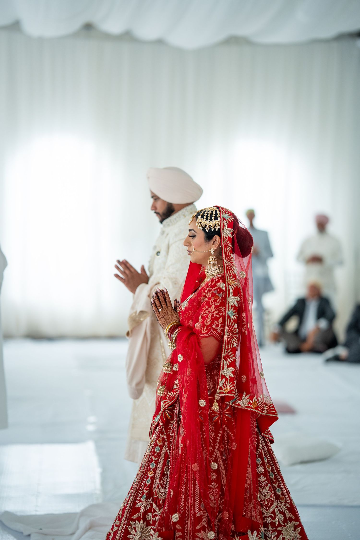 Wedding ceremony participants in traditional red and white attire praying in a temple setting.
