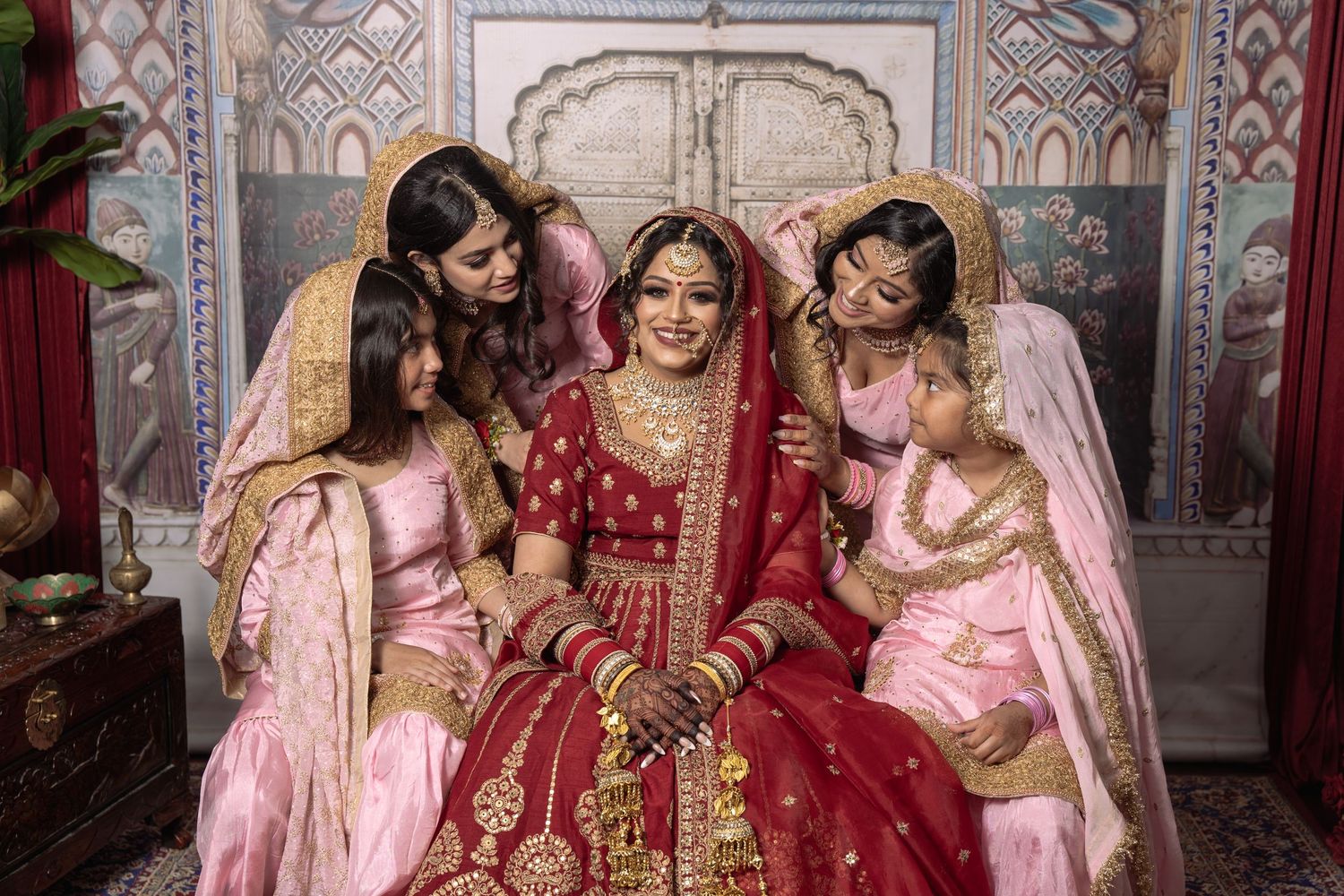 Wedding party in pink and red traditional Indian attire poses together against an ornate backdrop.