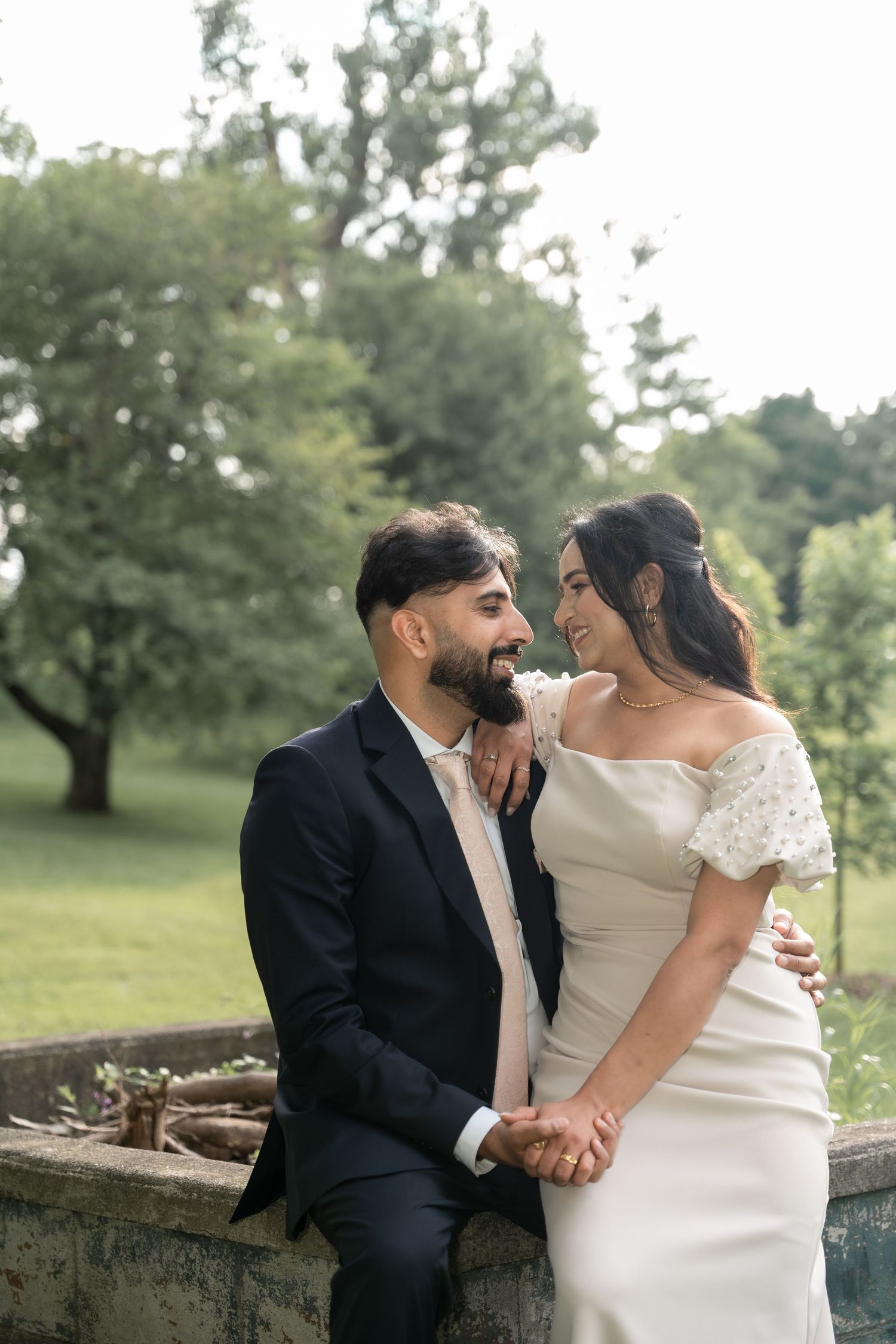 A romantic couple shares an intimate moment while sitting on a stone wall in a lush garden setting.