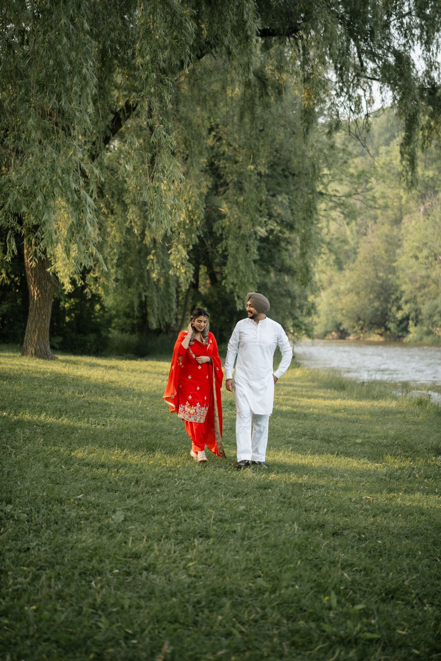 Couple walking through a park with willow trees, wearing traditional red and white Indian attire.