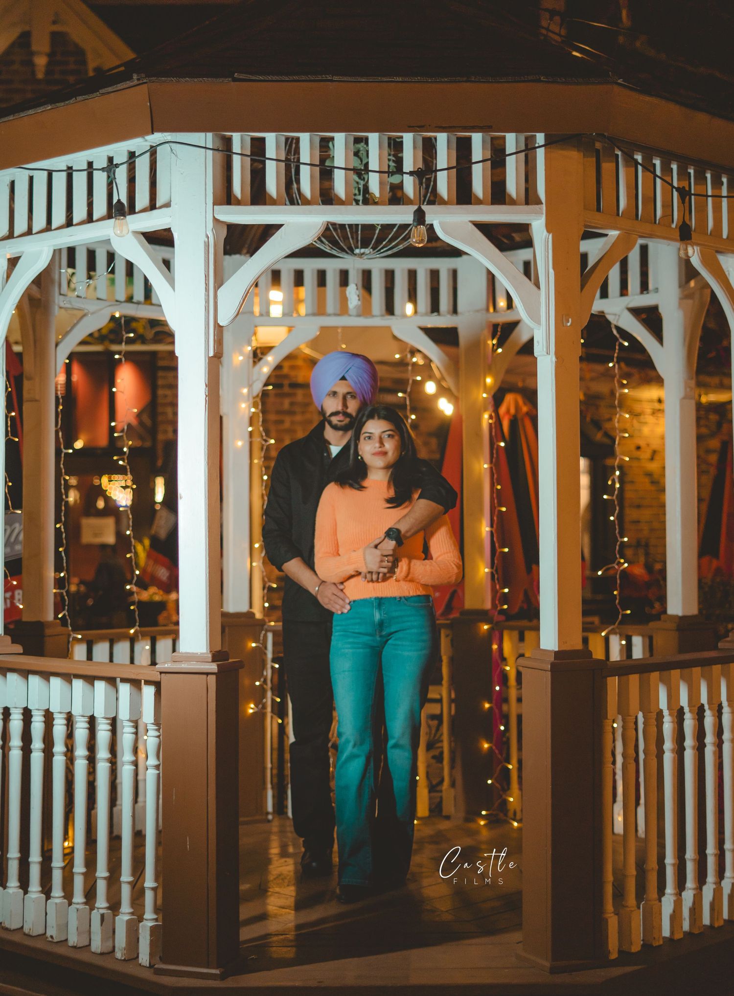 Romantic couple embraces in a beautifully lit white gazebo decorated with Christmas lights at night.