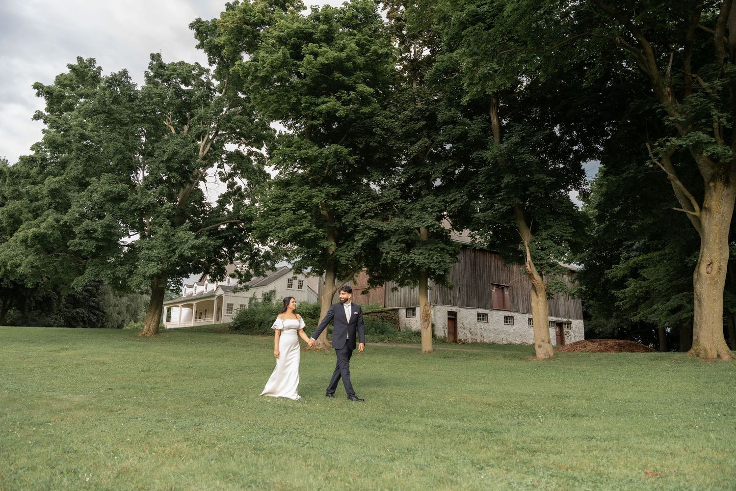 Wedding couple walking together on a lush green lawn surrounded by mature trees at a countryside venue.