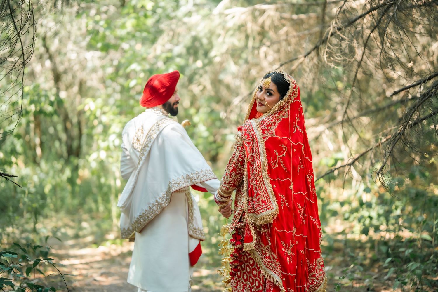 A Sikh bride in red lehenga and groom in white sherwani share a romantic moment on a forest path.