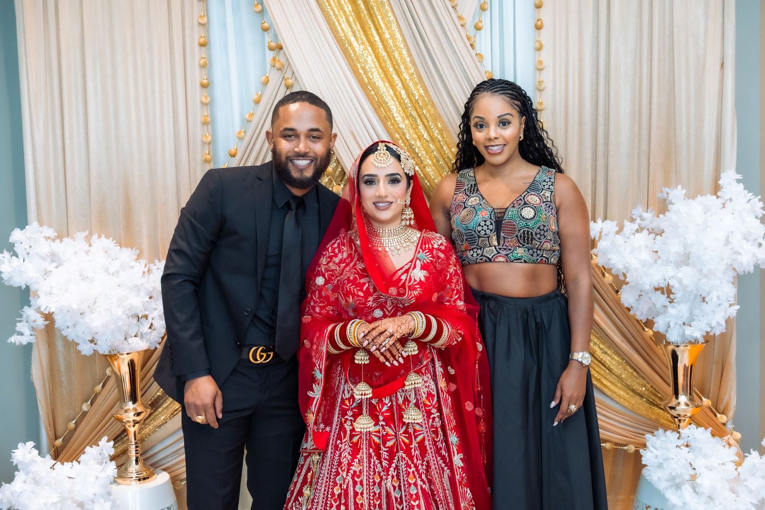 Wedding guests in formal attire pose together in front of elegant gold curtains and white floral decorations.
