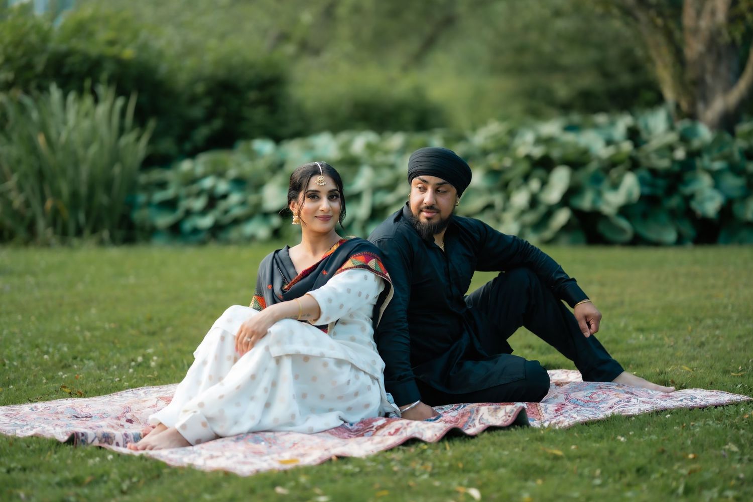 A couple relaxes on a picnic blanket in a lush green park setting during a sunny day.