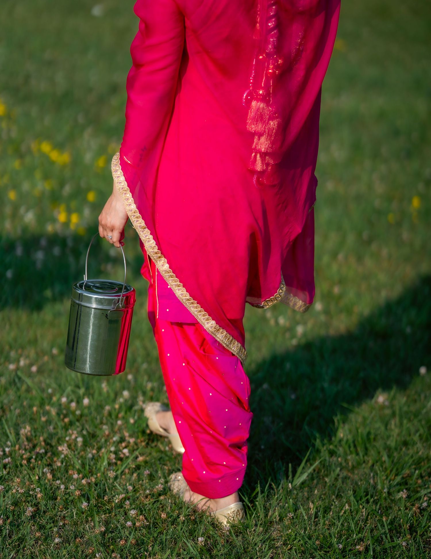A person wearing a bright pink traditional Indian dress carries a small metal container while walking on grass.