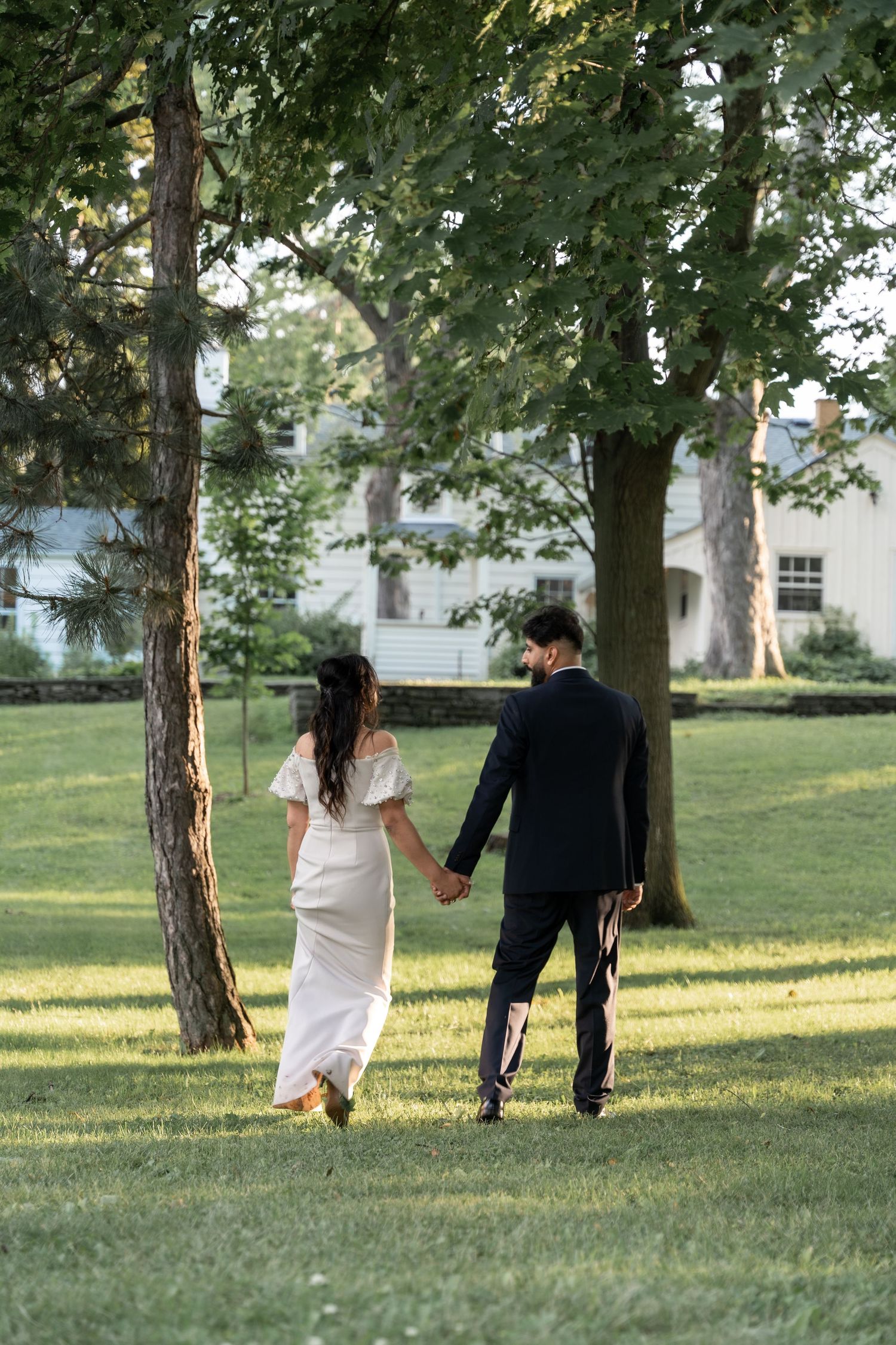 A couple in formal attire walks hand in hand through a sunlit garden with trees and a white building in the background.